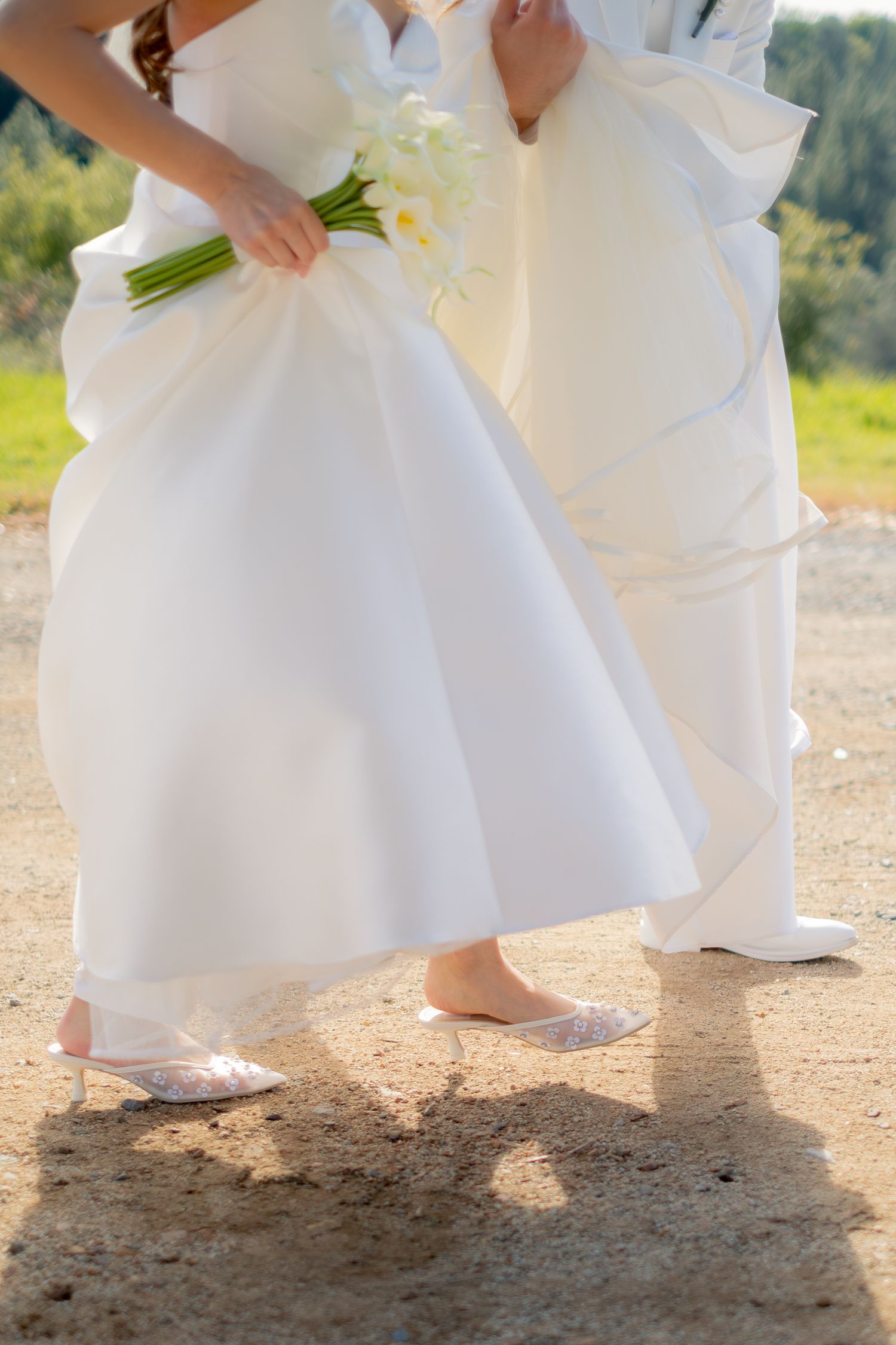 Close-up of bride and groom walking in white wedding attire, bride wearing lace flats holding calla lilies.