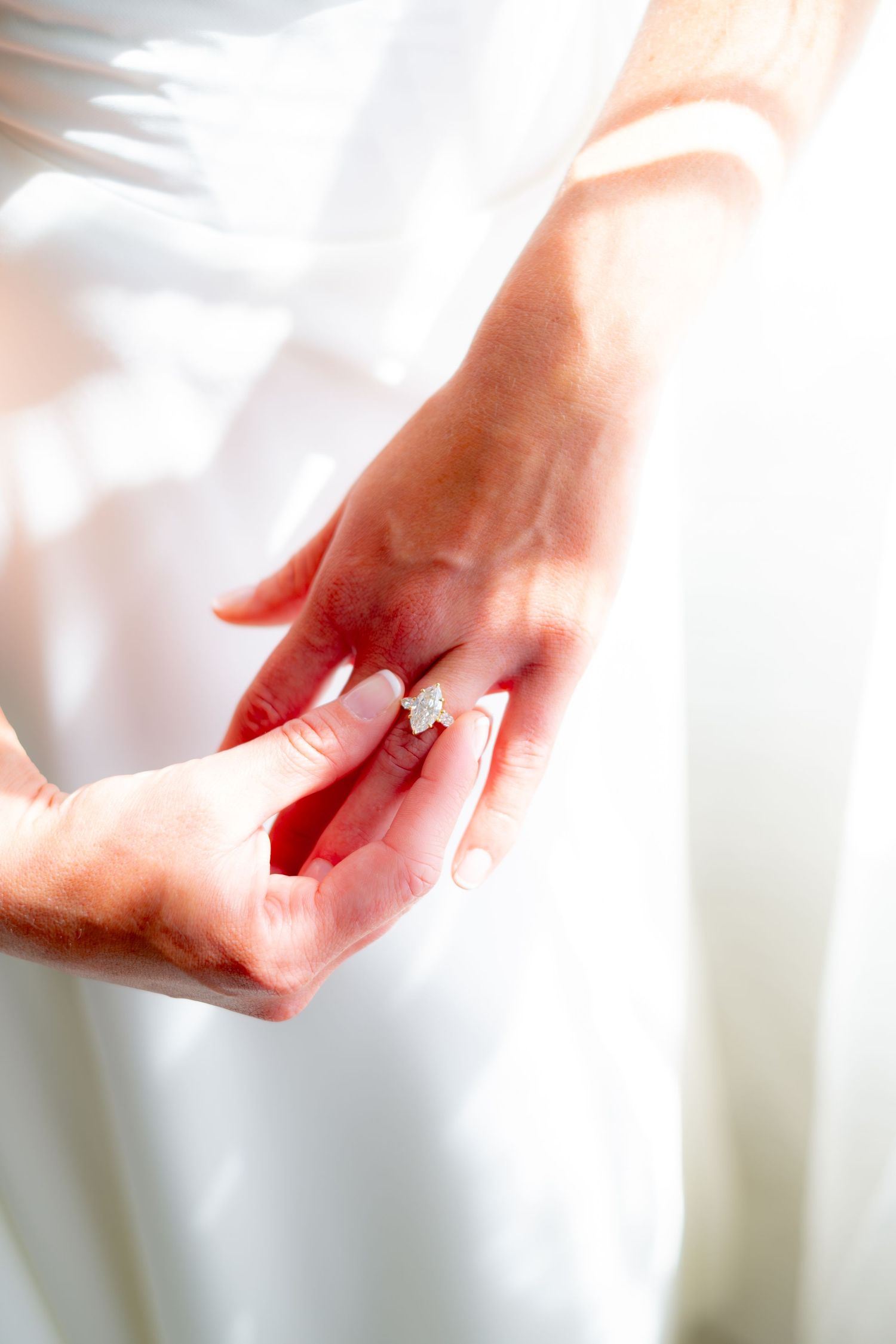 Close-up of hands placing a diamond engagement ring on a bride's finger against a white dress.