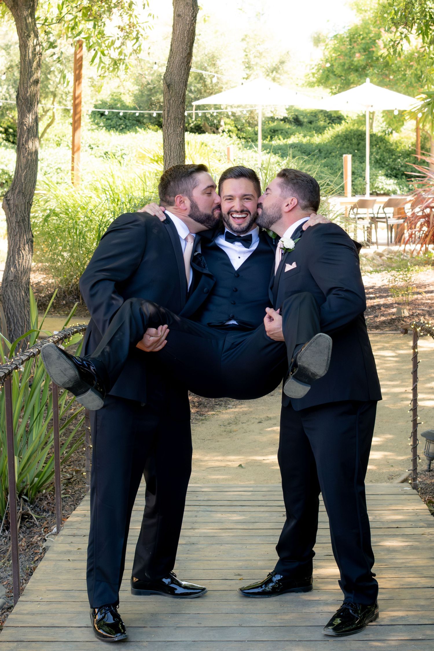Two groomsmen kiss and carry a laughing groom on a wooden bridge at an outdoor wedding venue.
