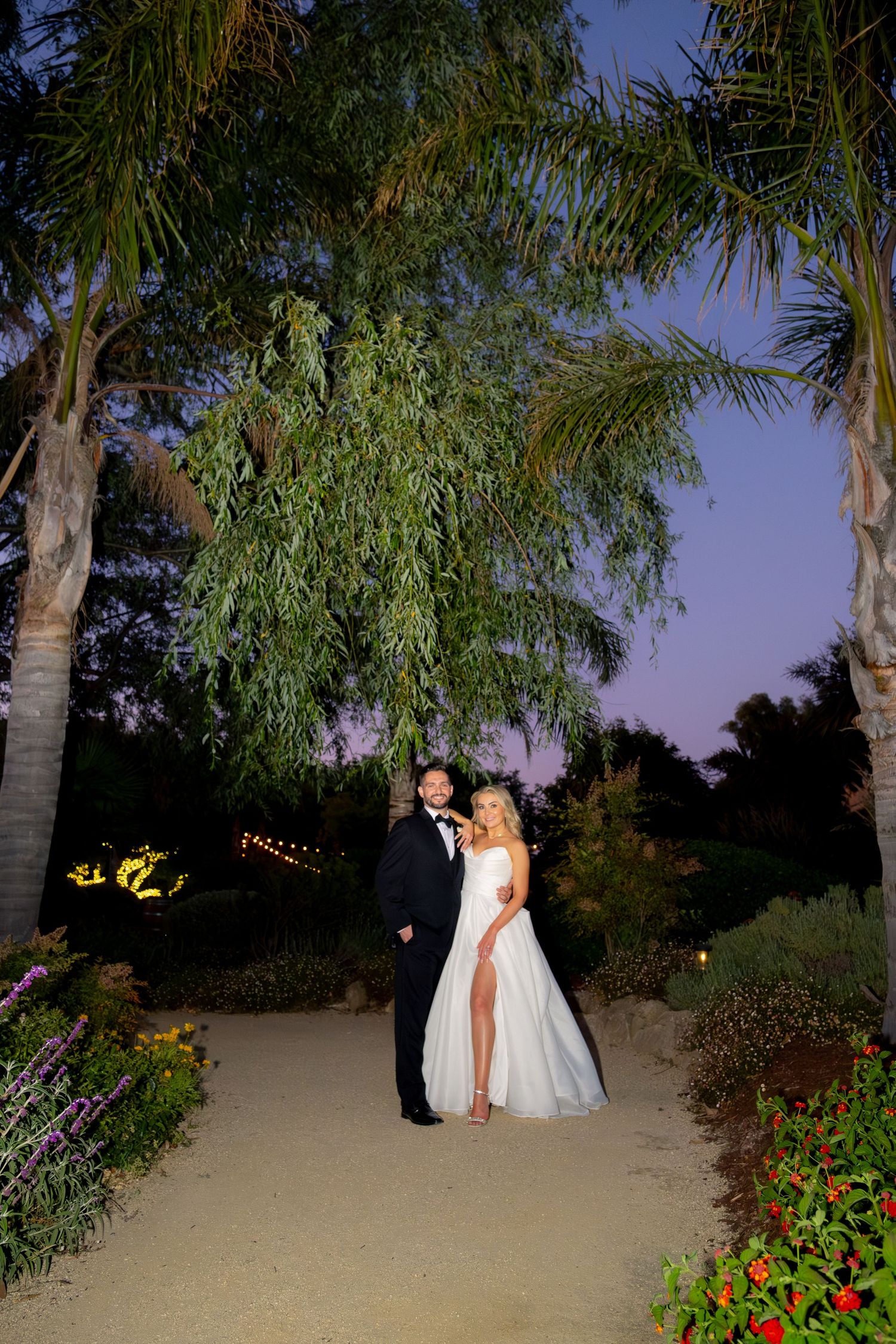 Bride and groom posing under palm trees at twilight during outdoor garden wedding reception.