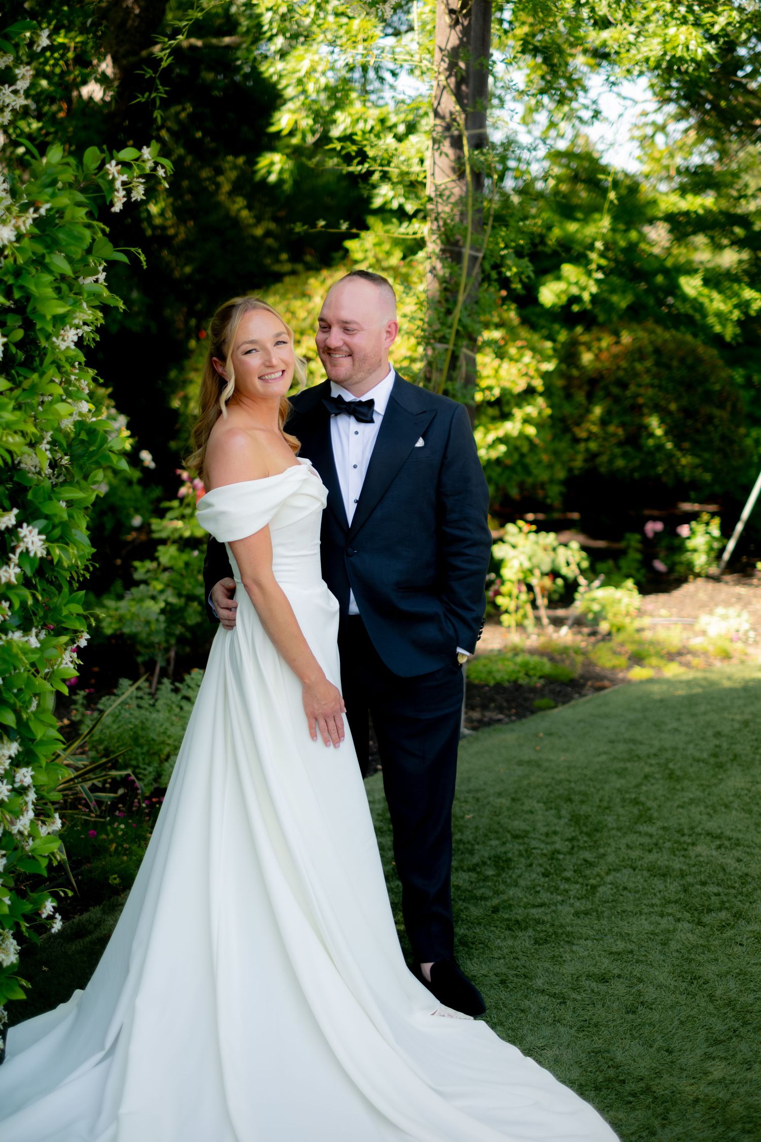 Bride in off-shoulder white gown and groom in black tuxedo posing together in a lush garden setting.