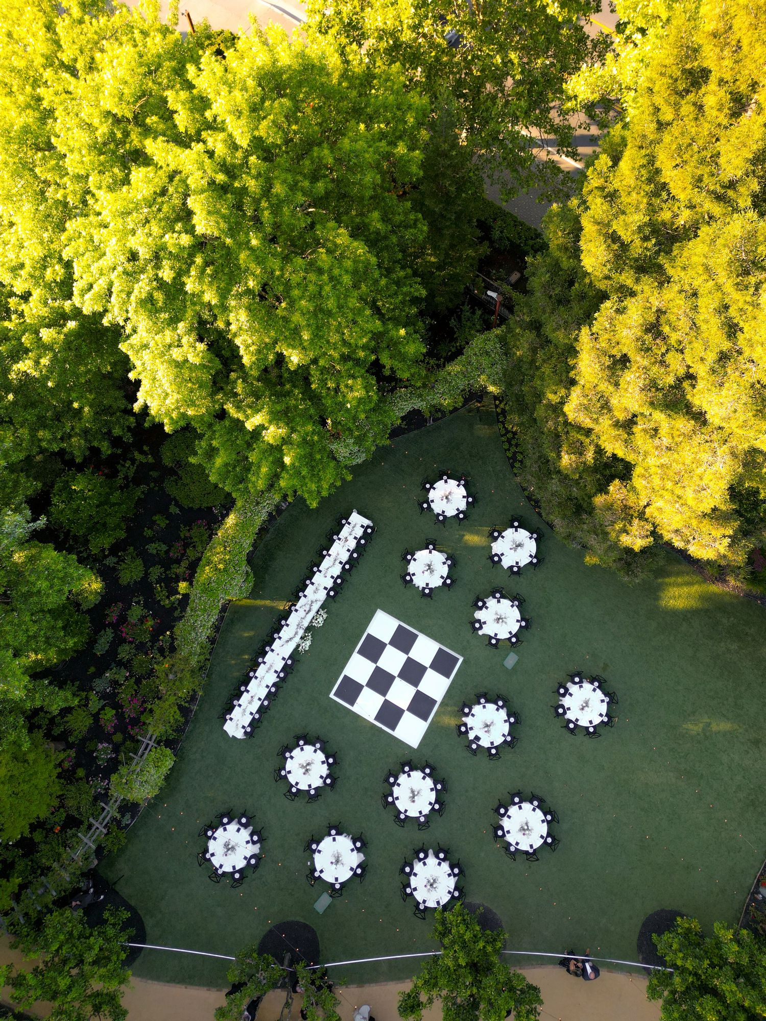 Aerial drone view of outdoor wedding reception with round white-draped tables surrounding a large checkerboard dance floor.