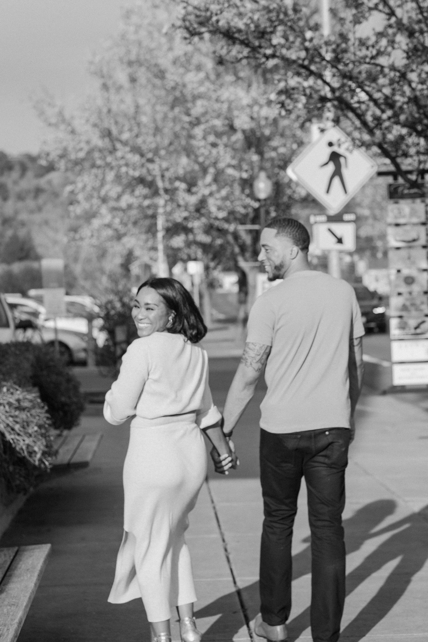 Black and white photo of a couple walking hand in hand down a city sidewalk together.