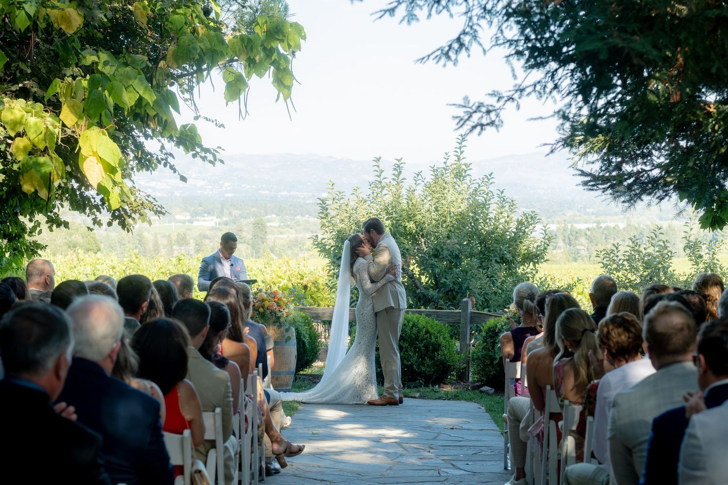 Couple shares first kiss at outdoor vineyard wedding ceremony with scenic mountain valley views behind them.