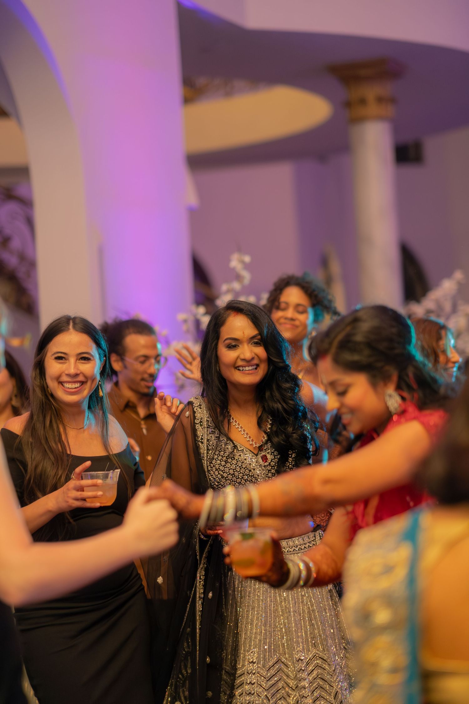 Group of women in Indian and western attire smiling and dancing together at a vibrant wedding reception.