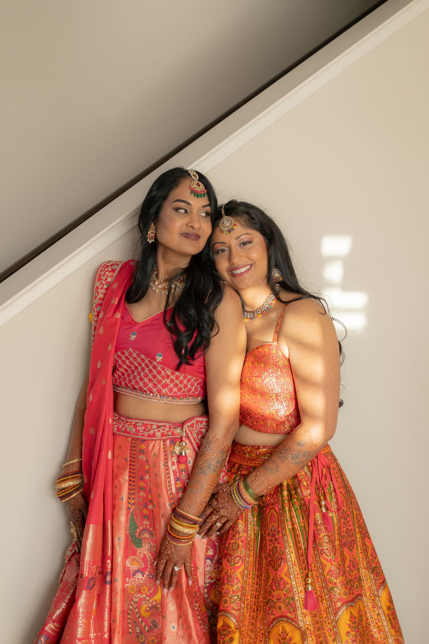 Two young women in vibrant Indian lehenga outfits with gold jewelry pose together on a staircase.