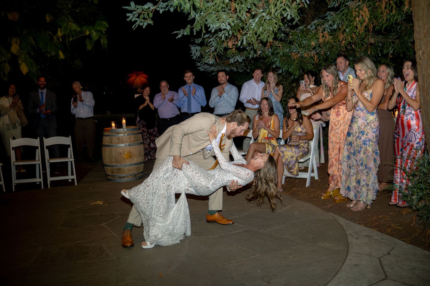 Groom dips bride dramatically on dance floor surrounded by cheering wedding guests at night.