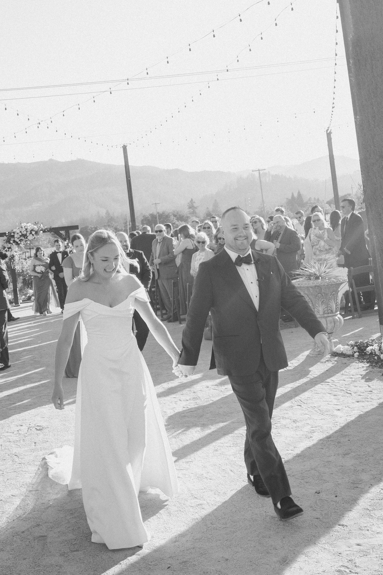 Newlyweds joyfully walk hand in hand at an outdoor vineyard wedding reception in black and white.