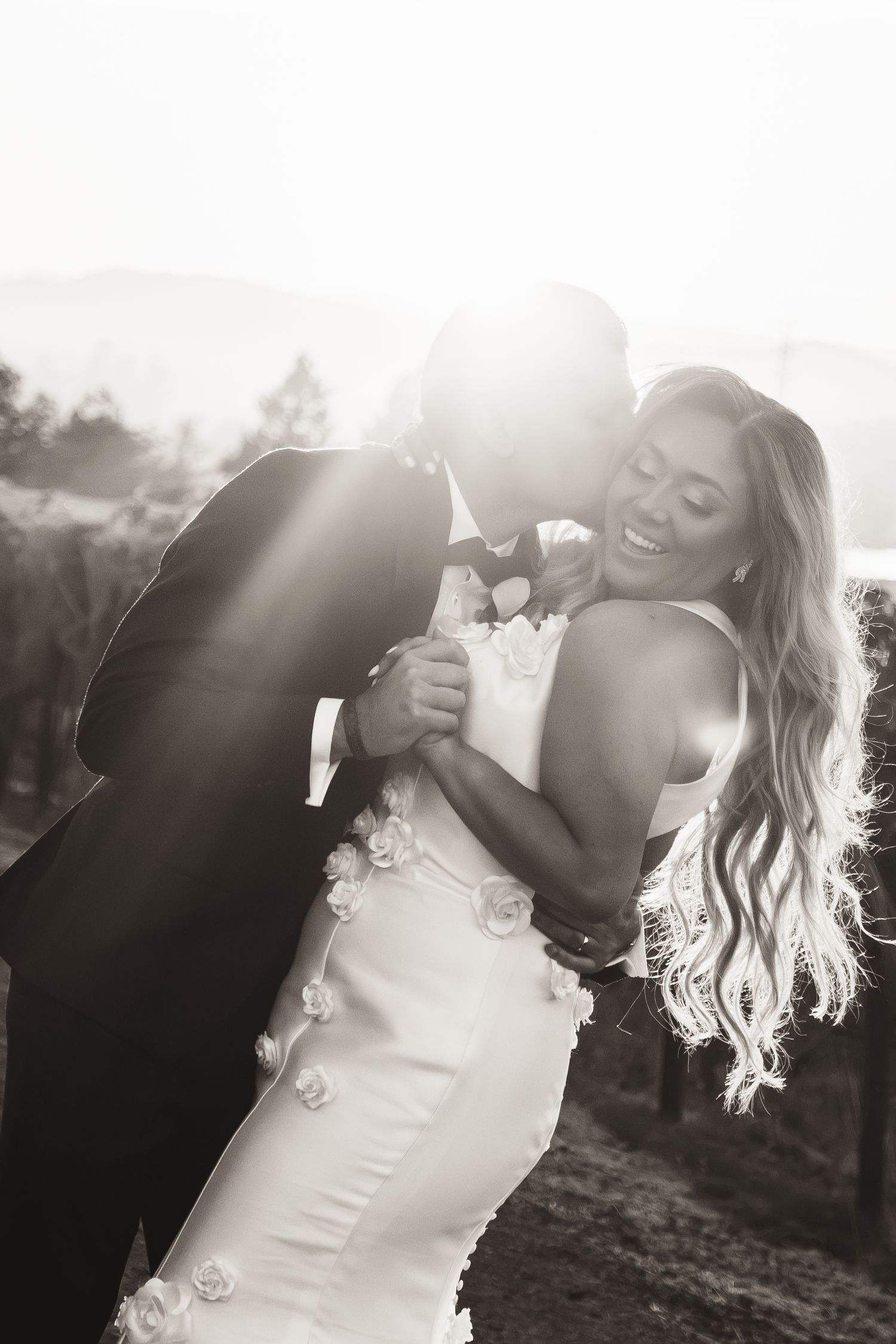 Black and white photo of groom kissing laughing bride in a vineyard with dramatic backlit sunshine.