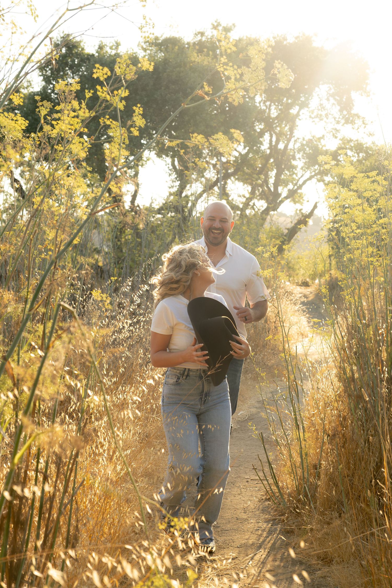Happy couple walking together on a sunlit path surrounded by golden wildflowers and trees.