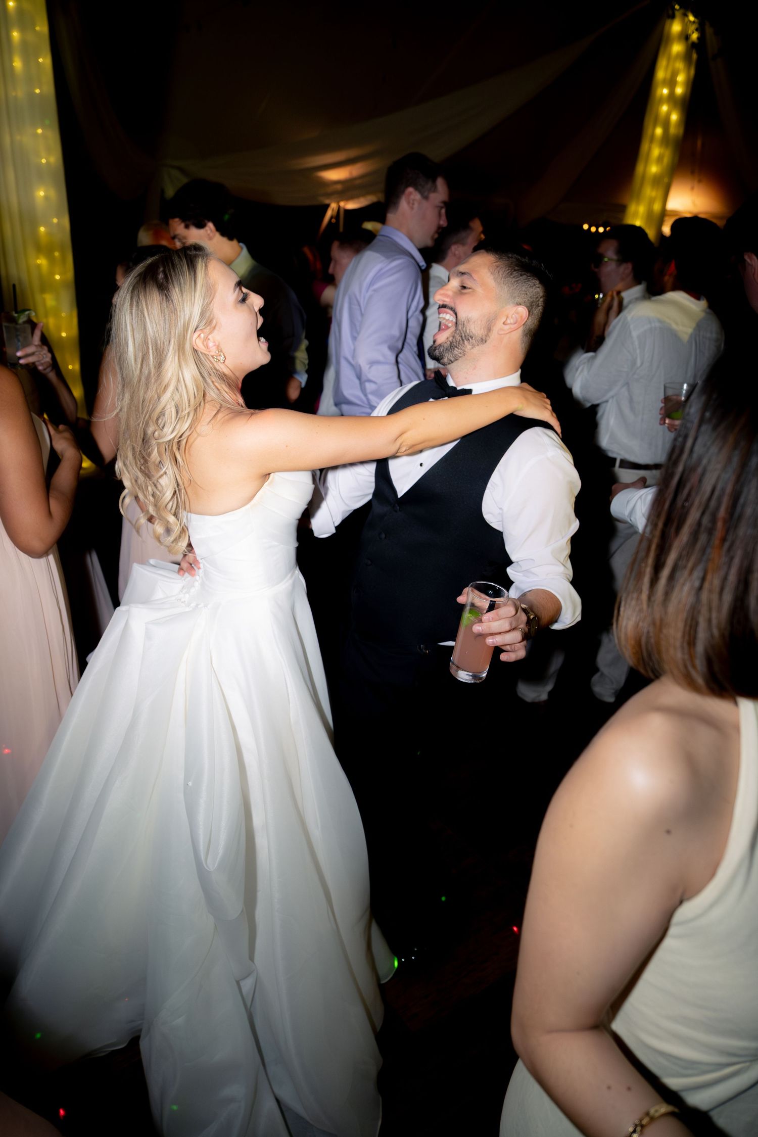 Bride in white gown and groom in vest share a joyful dance at wedding reception.
