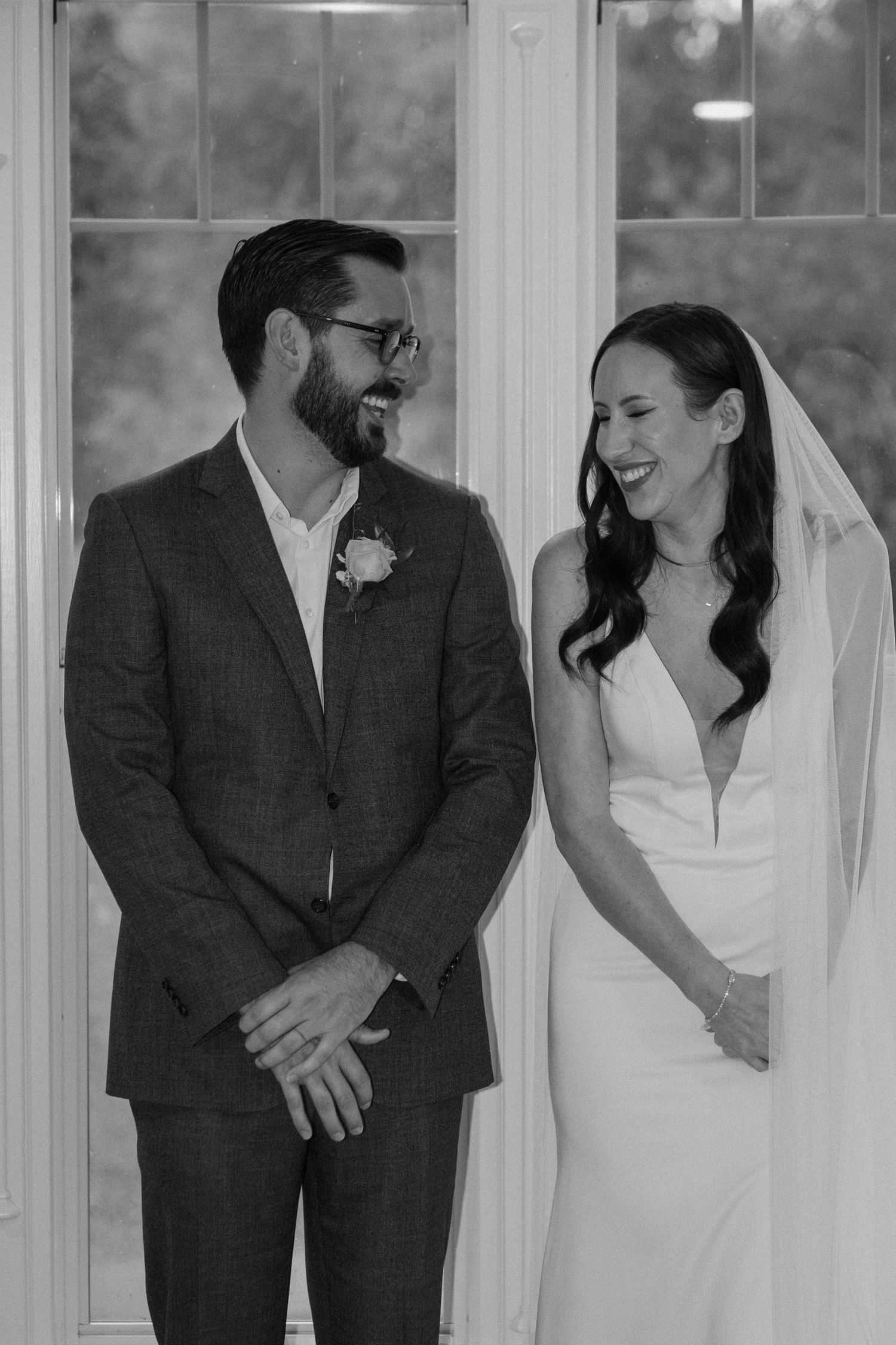 Laughing bride and bearded groom in gray suit share a joyful moment in black and white wedding photo.