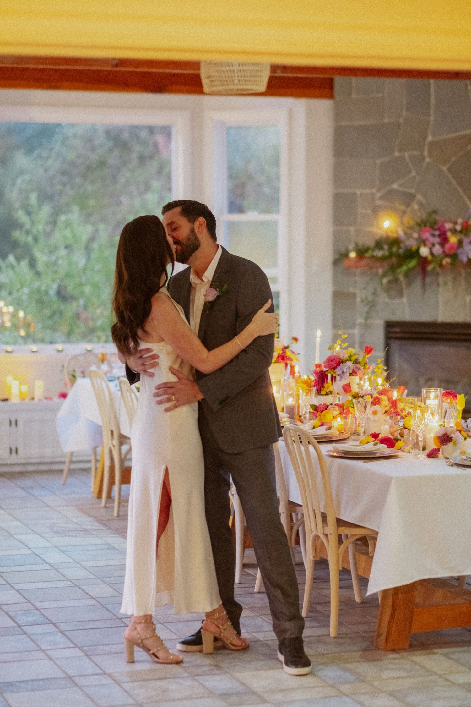 Bride and groom share a romantic kiss beside an elegantly decorated wedding reception table with colorful floral arrangements.