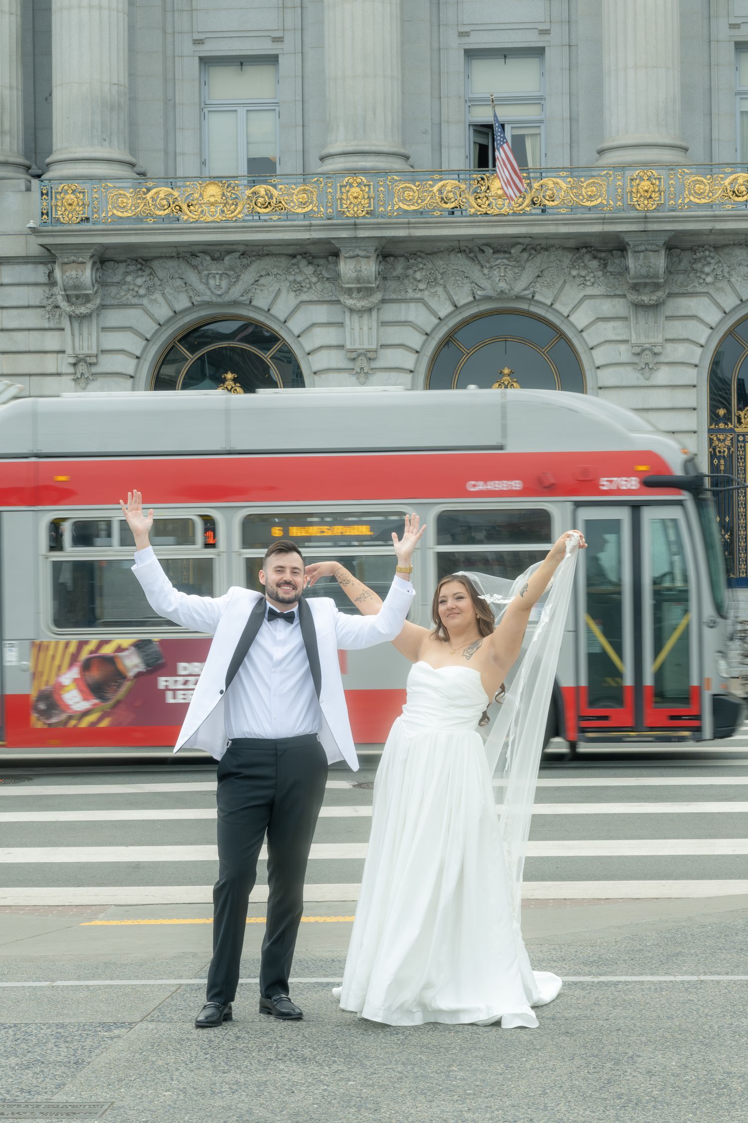 Joyful bride and groom raising arms on crosswalk with red city bus behind them.