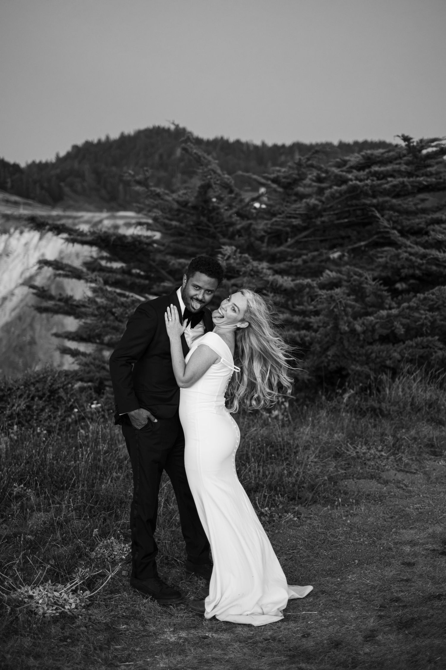 Black and white photo of laughing bride and groom embracing on a windswept hillside with pine trees.