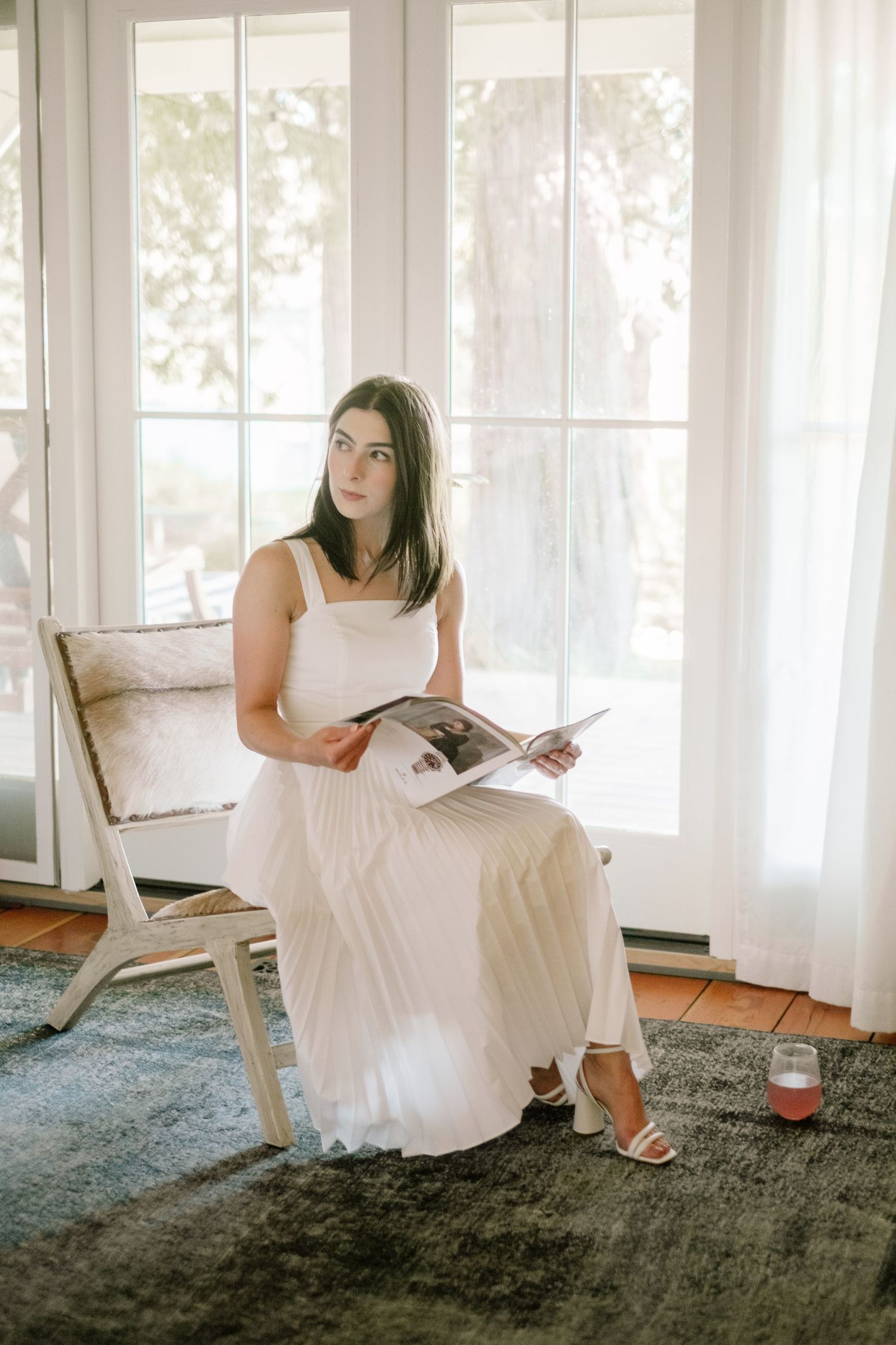 Bride in white pleated dress reads magazine while seated by sunlit French doors with sheer curtains.