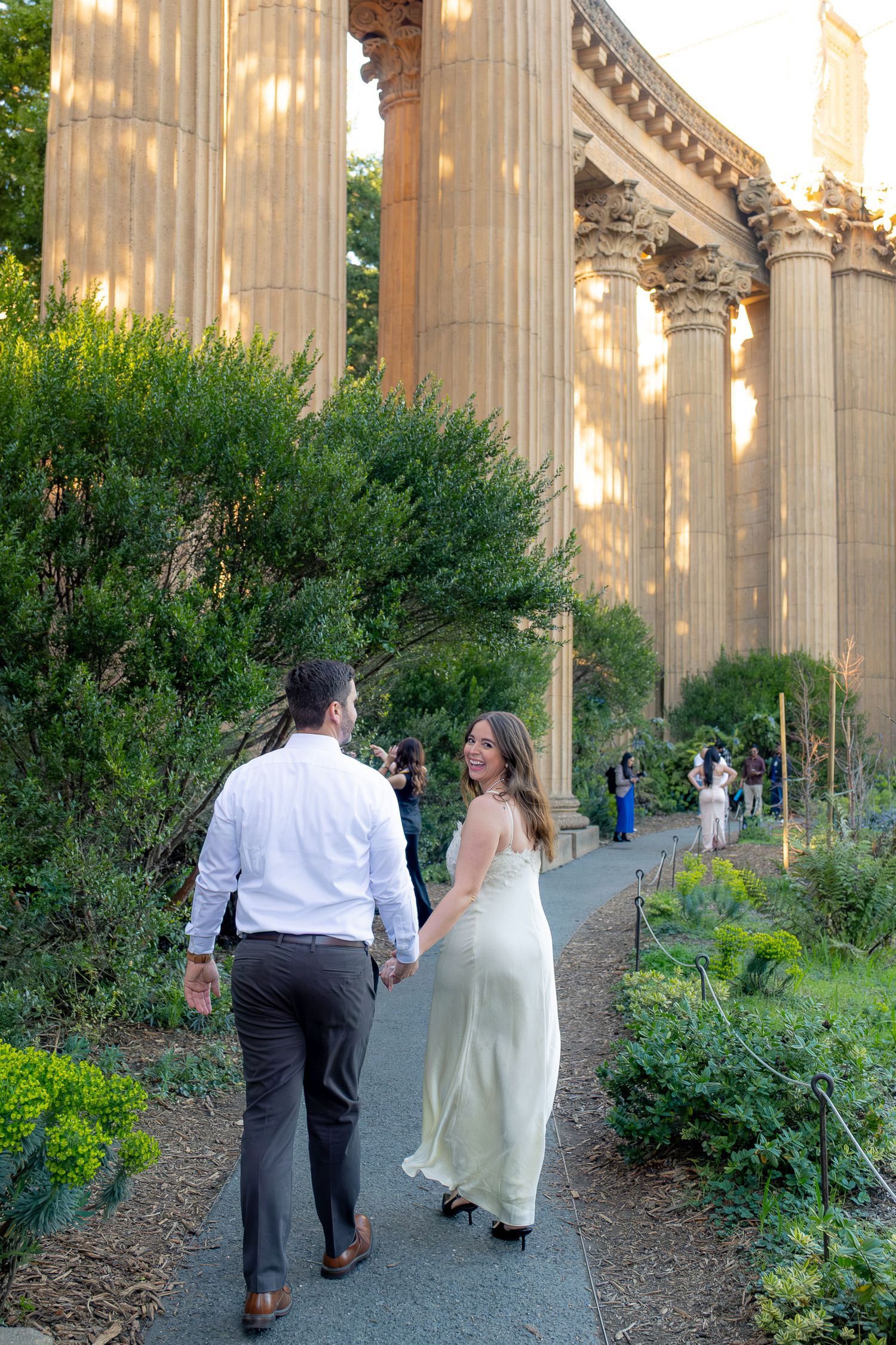 Couple in wedding attire holding hands while walking past grand columns at the Palace of Fine Arts in San Francisco.