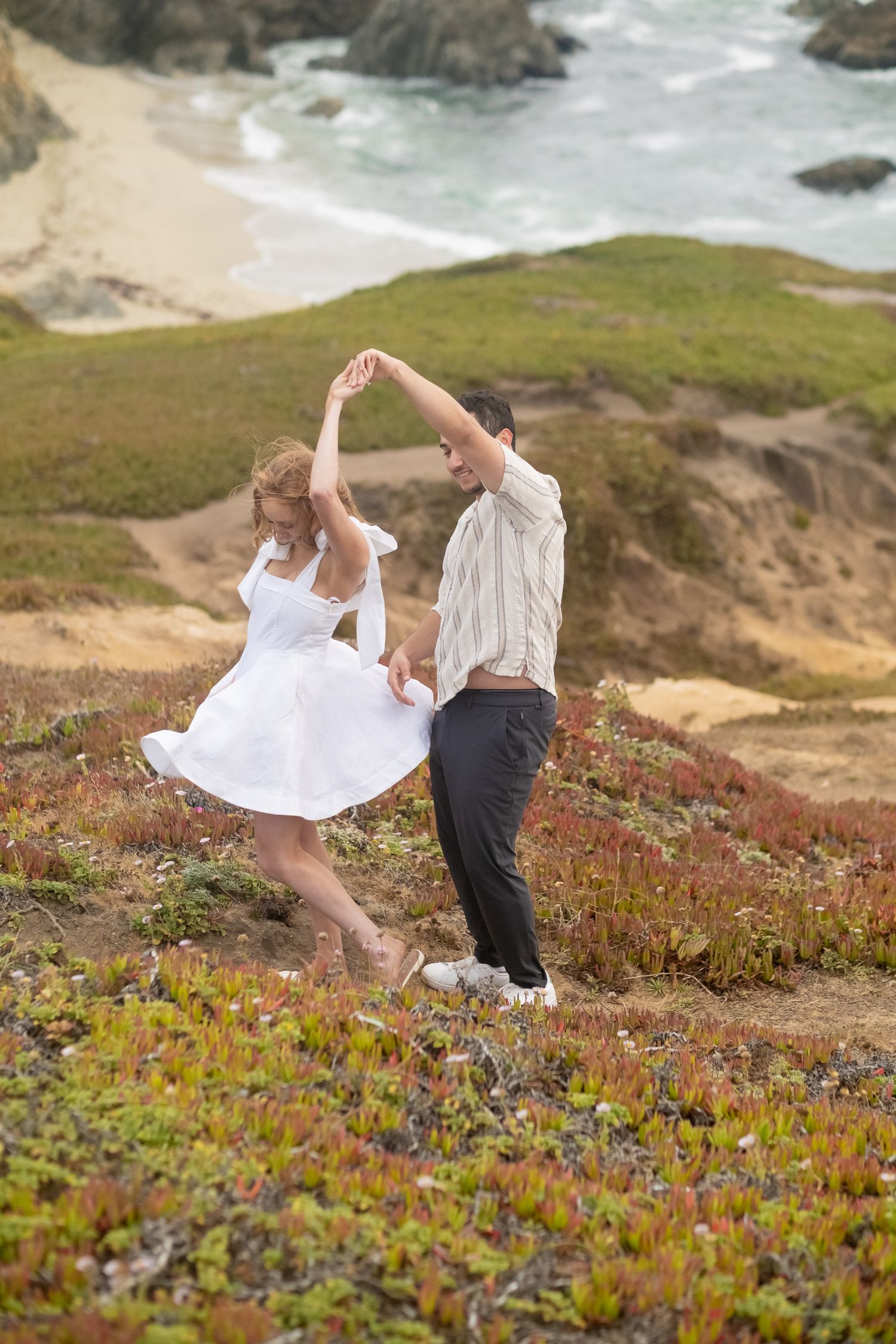 Couple dancing joyfully on coastal bluff with ocean view, man spinning woman in white dress outdoors.