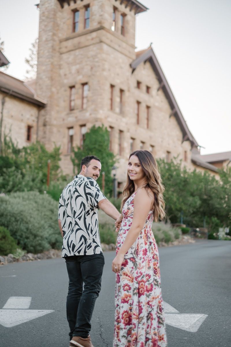 Couple holding hands walking near a stone castle building, woman smiling over shoulder.