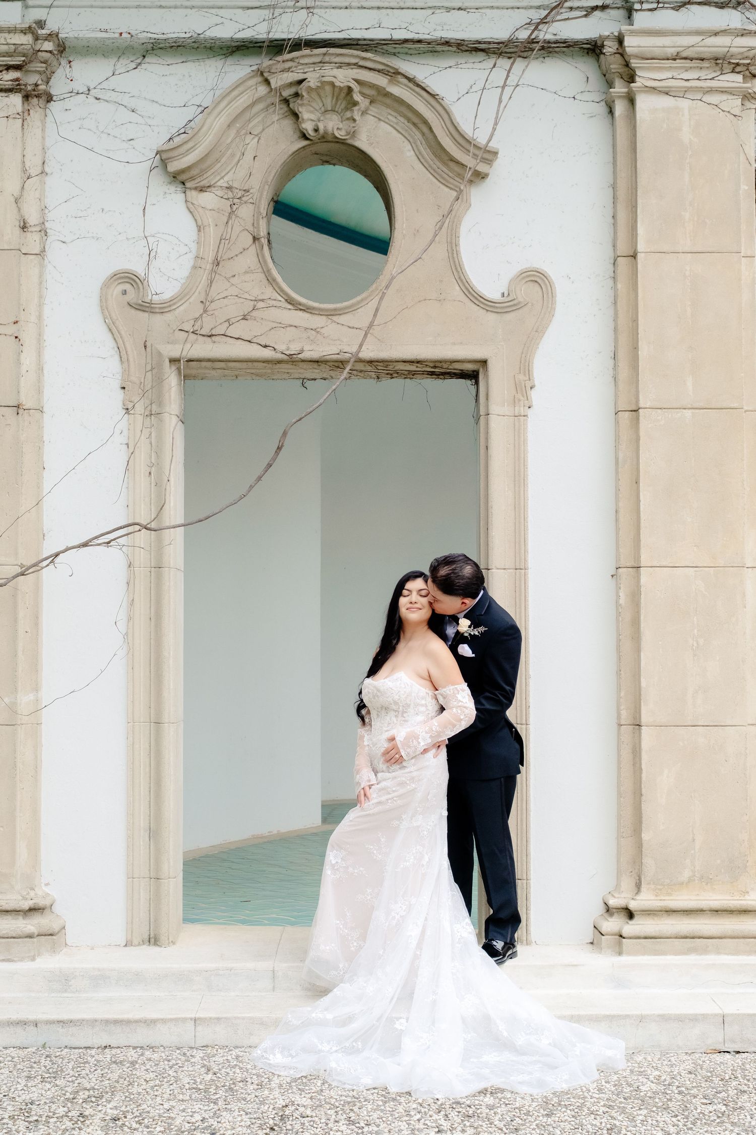 Bride in lace mermaid gown and groom share romantic kiss in ornate arched doorway of classical building.