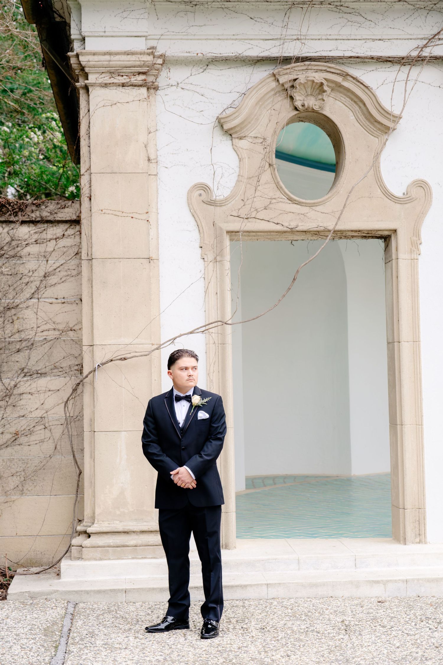 Groom in black tuxedo with bow tie and boutonniere standing before ornate stone archway at wedding venue.