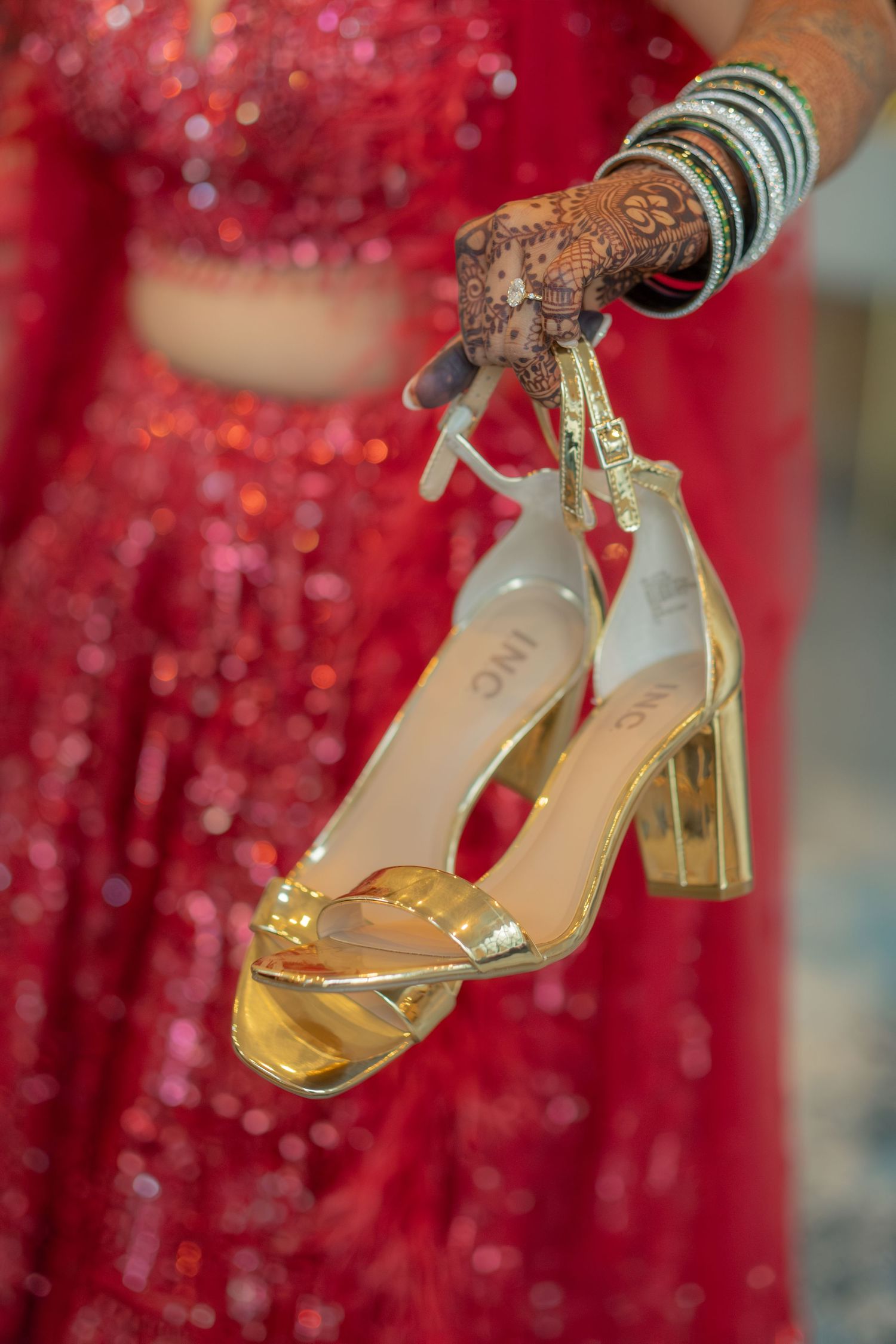 Bride in red lehenga with henna-adorned hands holding gold block heel sandals at Indian wedding.