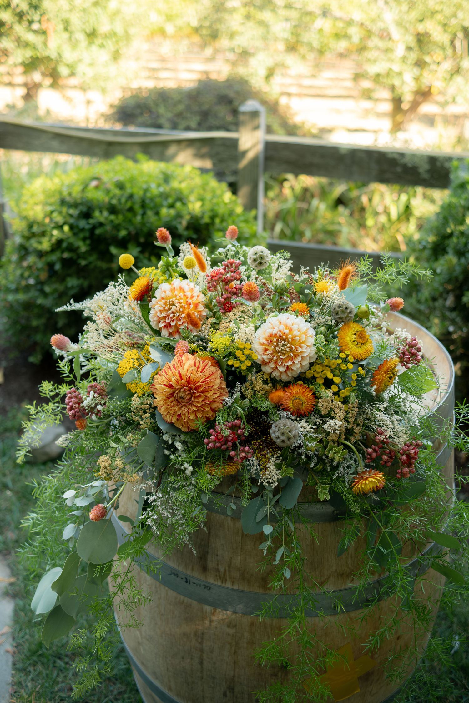 Rustic wooden barrel topped with a lush floral arrangement of orange dahlias and wildflowers outdoors.