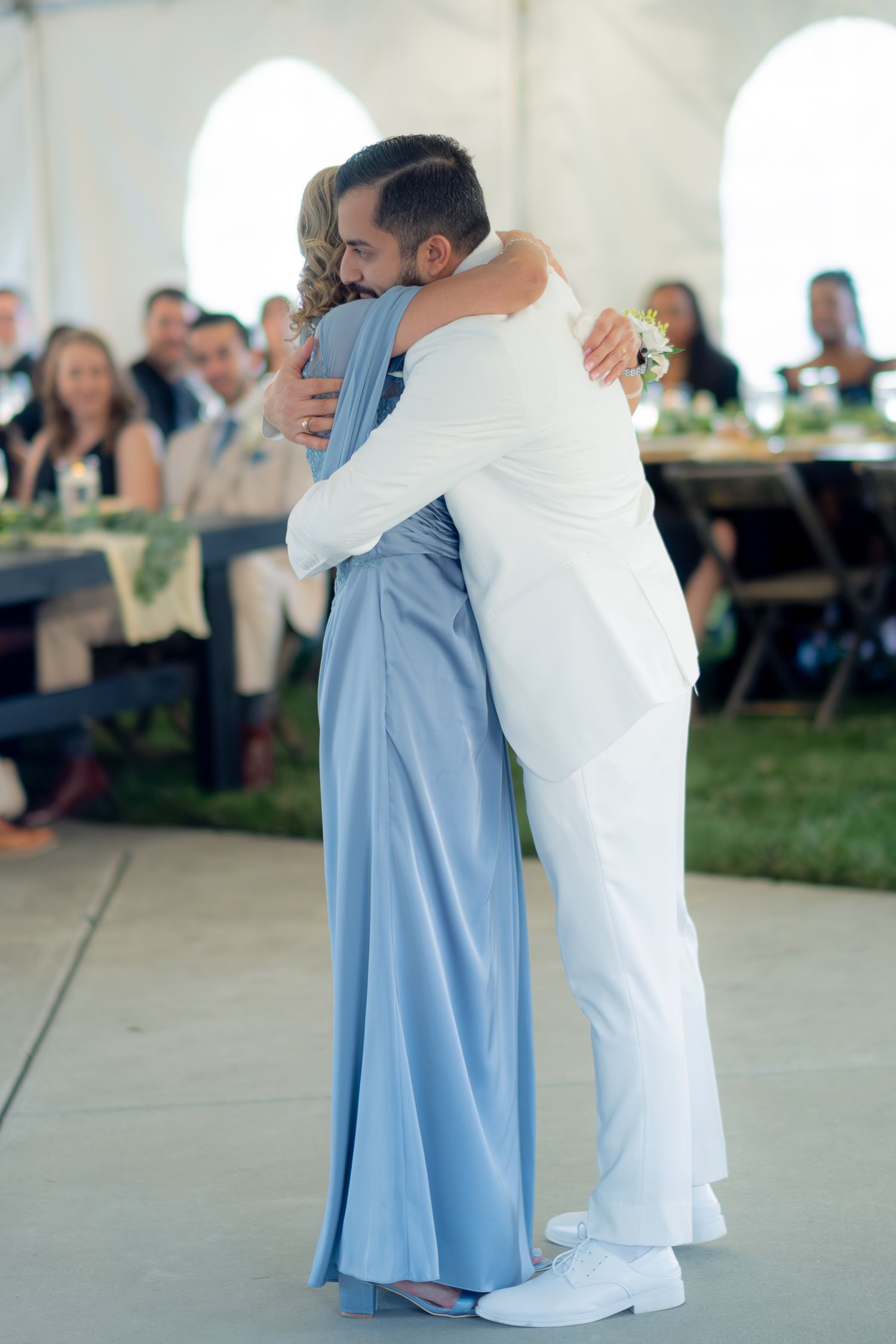 Groom in white suit sharing emotional dance and hug with mother in blue gown at tented wedding reception.