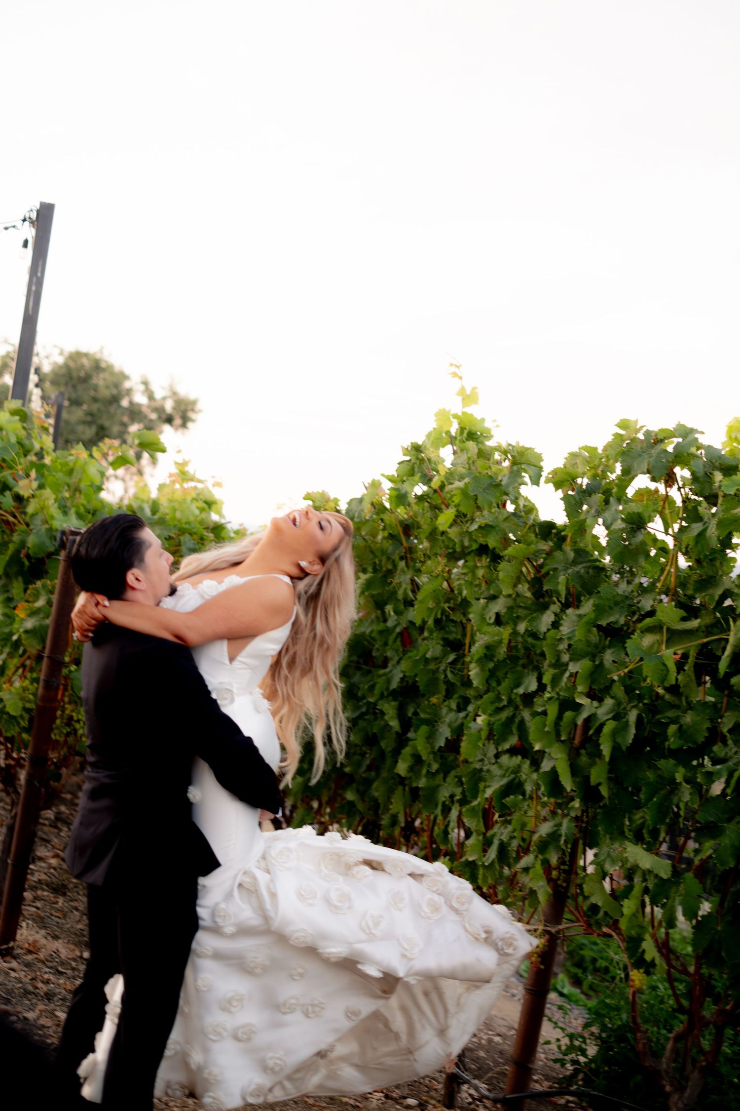 Groom lifts smiling bride in white mermaid gown among vineyard rows at outdoor wedding.