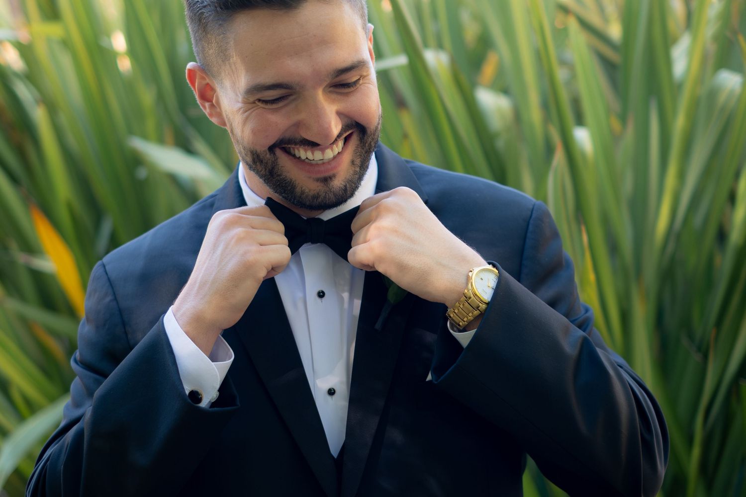 Handsome groom in black tuxedo and bow tie smiling while adjusting his collar outdoors.