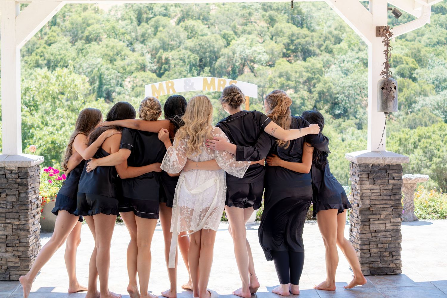 Bride and bridesmaids in black stand arm-in-arm on an outdoor pavilion overlooking lush green trees.