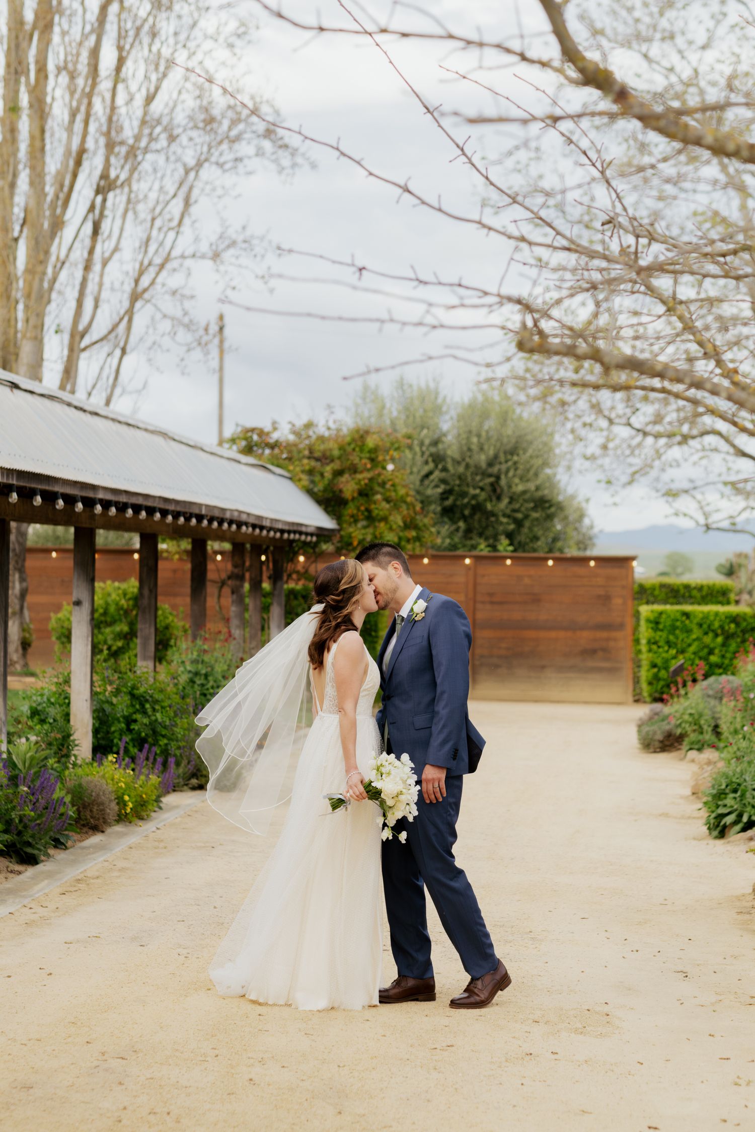 Bride and groom share a romantic kiss on a garden path, bride in white gown with veil, groom in navy suit.