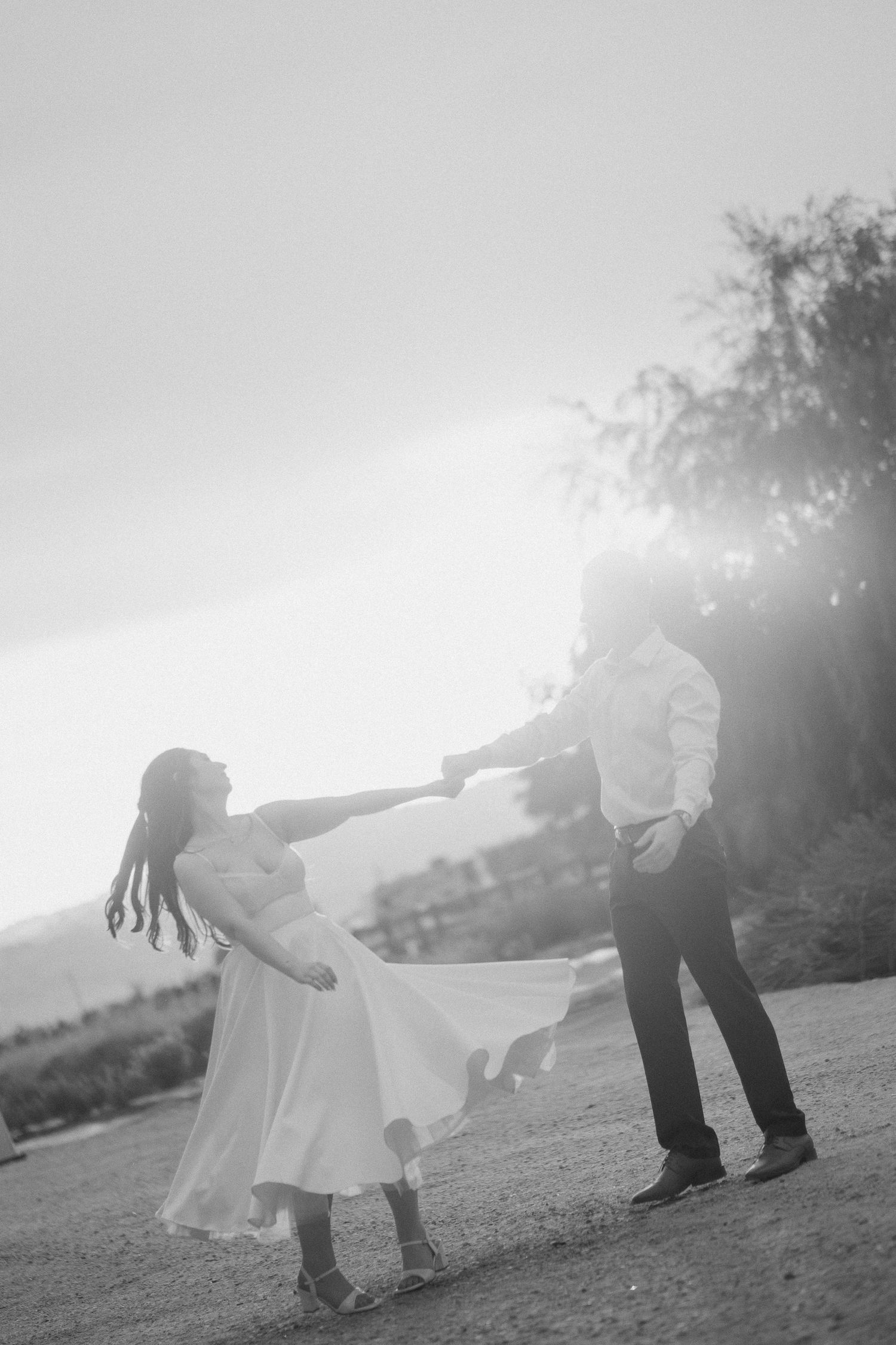 Black and white photo of a couple dancing outdoors at sunset, woman in white dress twirling while holding hands with man.