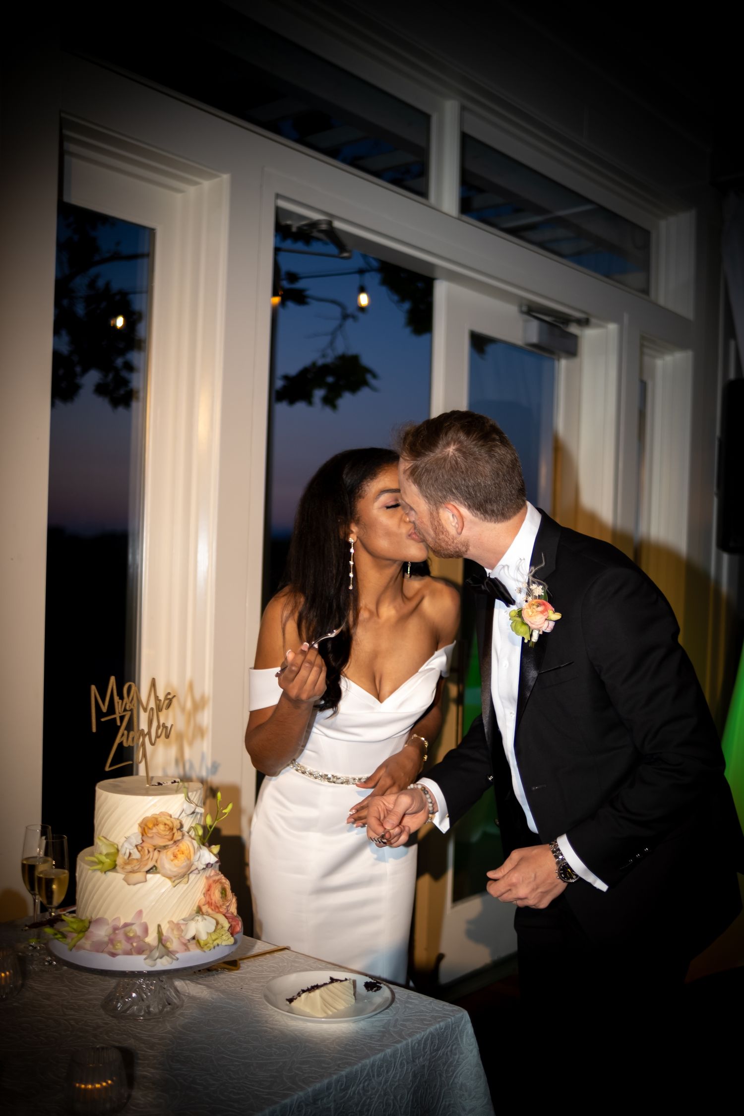 Bride and groom share a kiss while cutting their decorated wedding cake at evening reception.