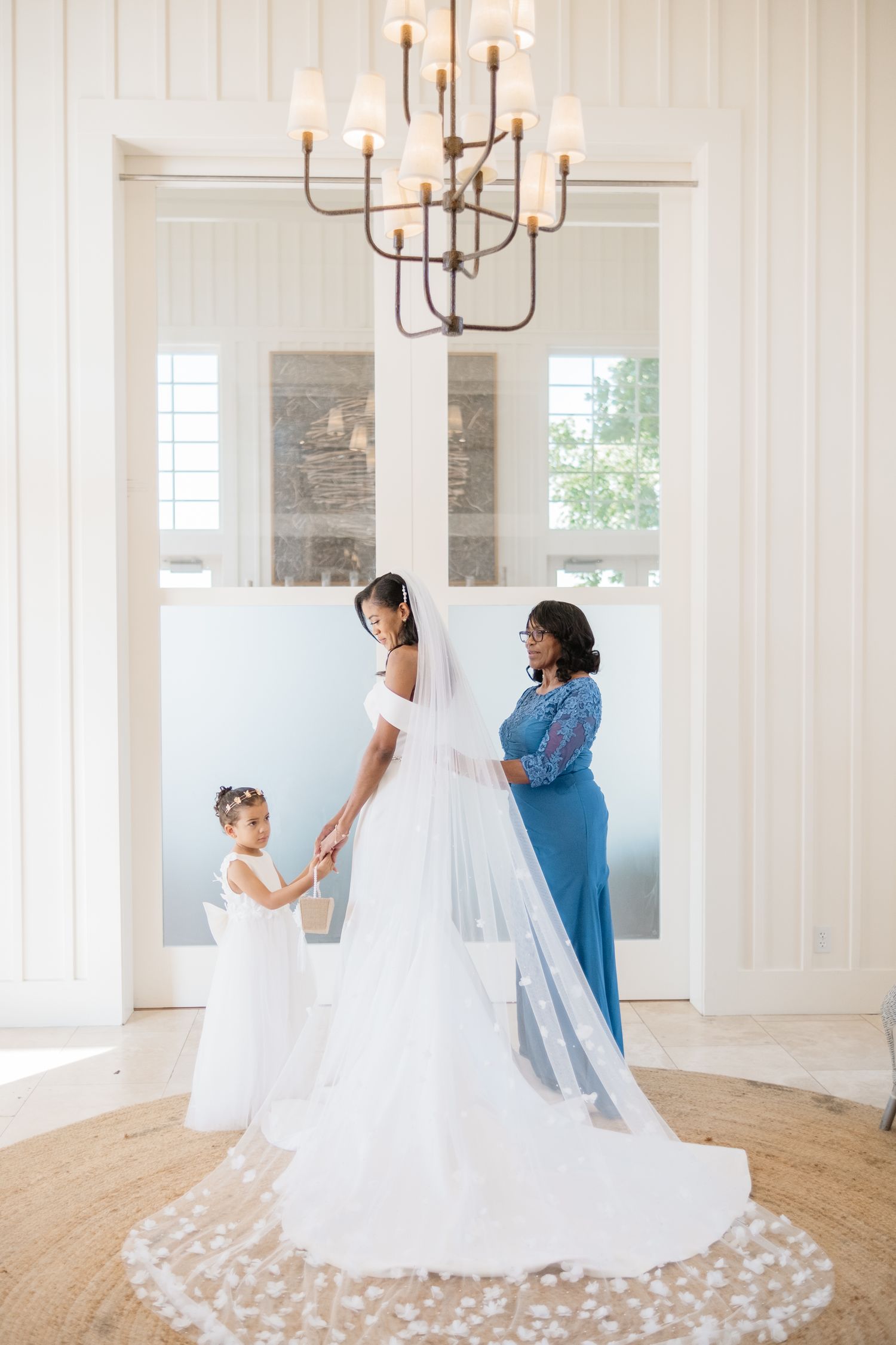 Bride in white off-shoulder gown with long veil stands with flower girl and woman in blue dress under chandelier.