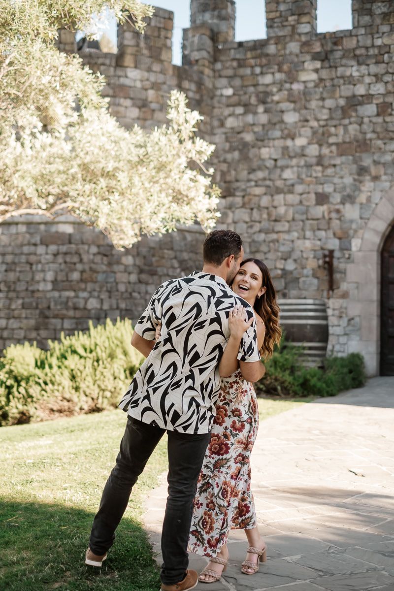 A laughing couple dances outside a stone castle, woman in floral skirt, man in black and white patterned shirt.