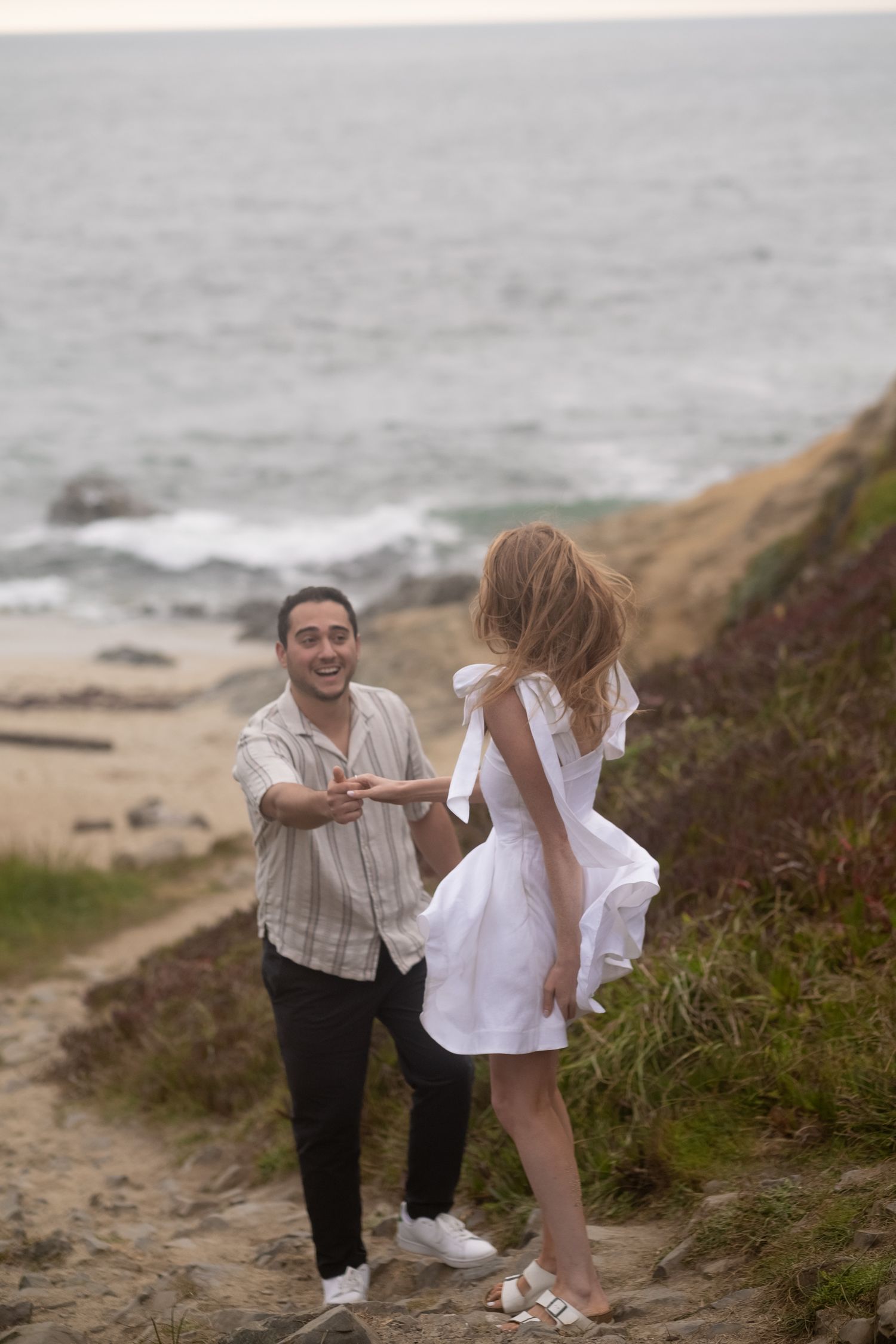 Man laughing and reaching toward woman in white dress on a scenic coastal hillside path.