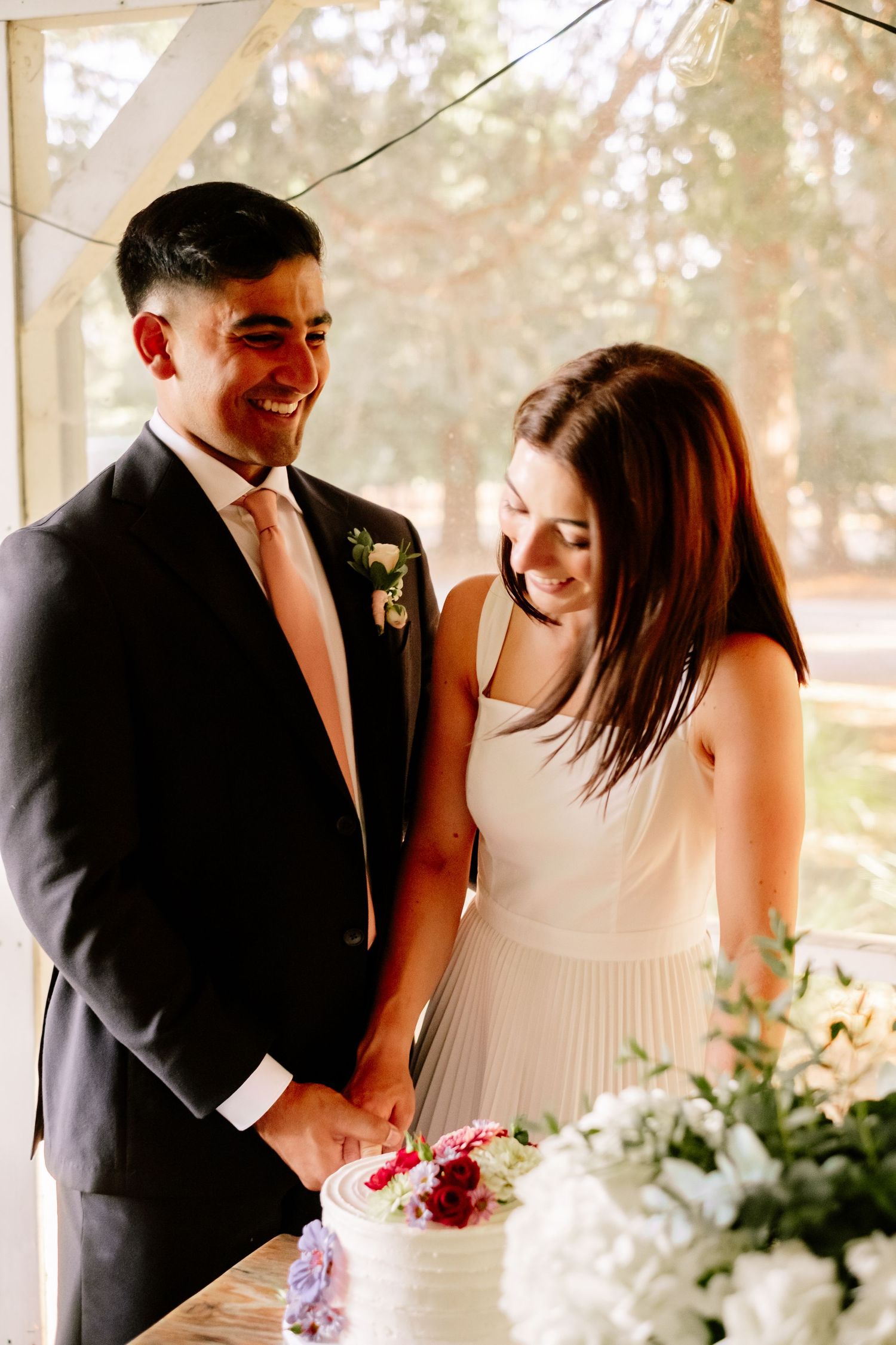 Smiling bride and groom cutting their white wedding cake decorated with colorful flowers.
