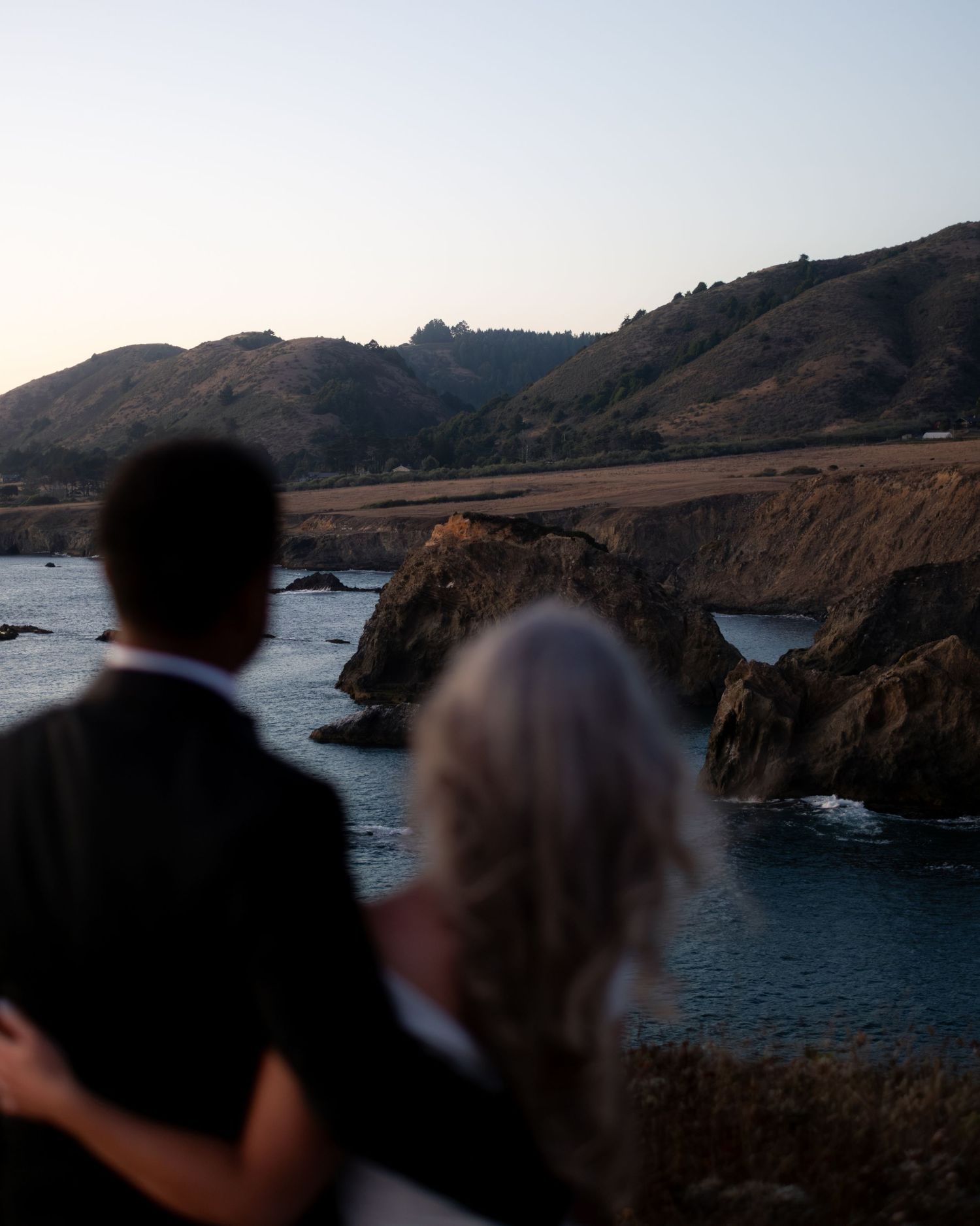 Couple embracing while overlooking scenic California coastal cliffs and ocean at dusk.