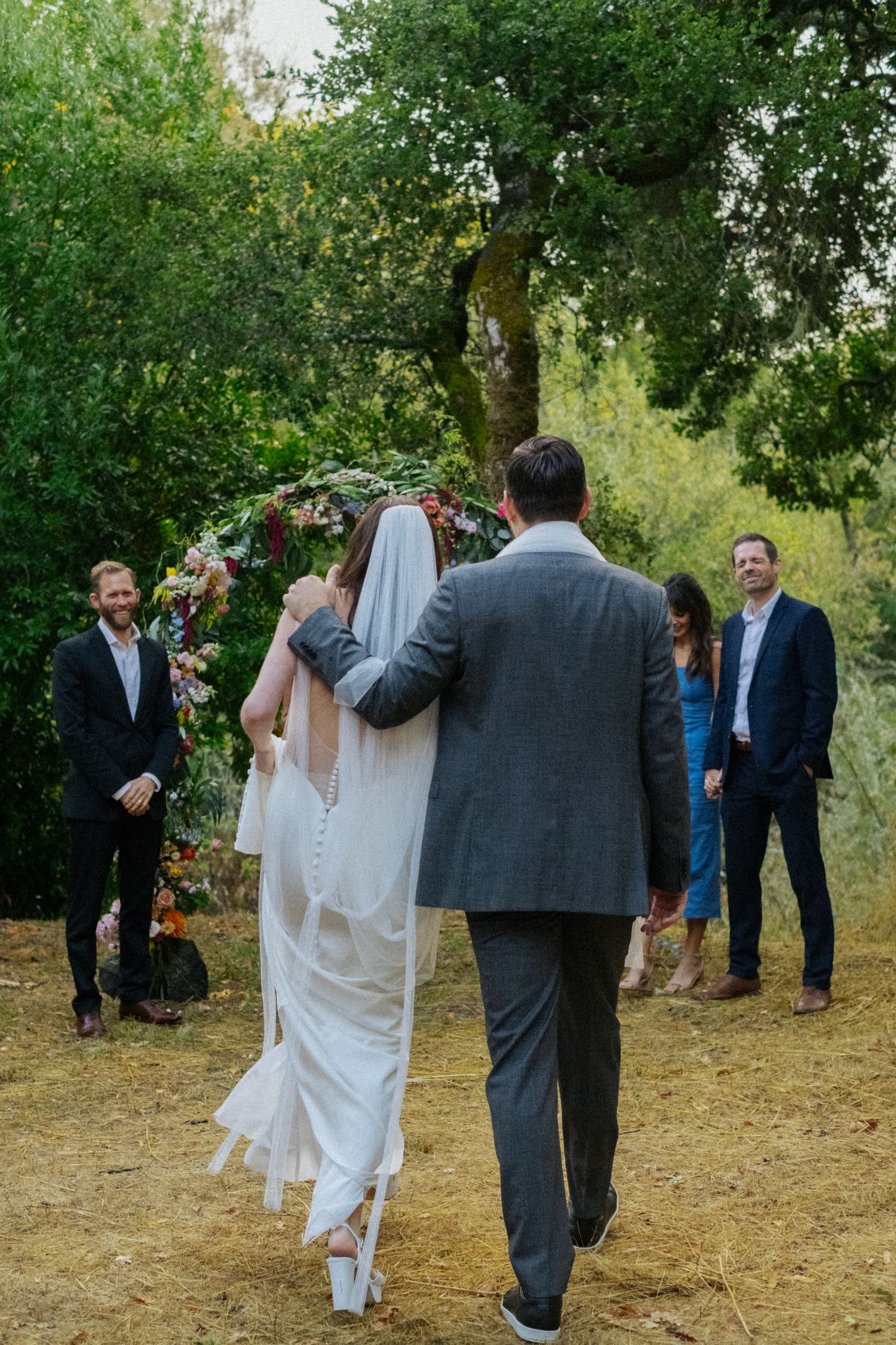 Bride and groom walking toward floral arch during outdoor wedding ceremony with guests watching.
