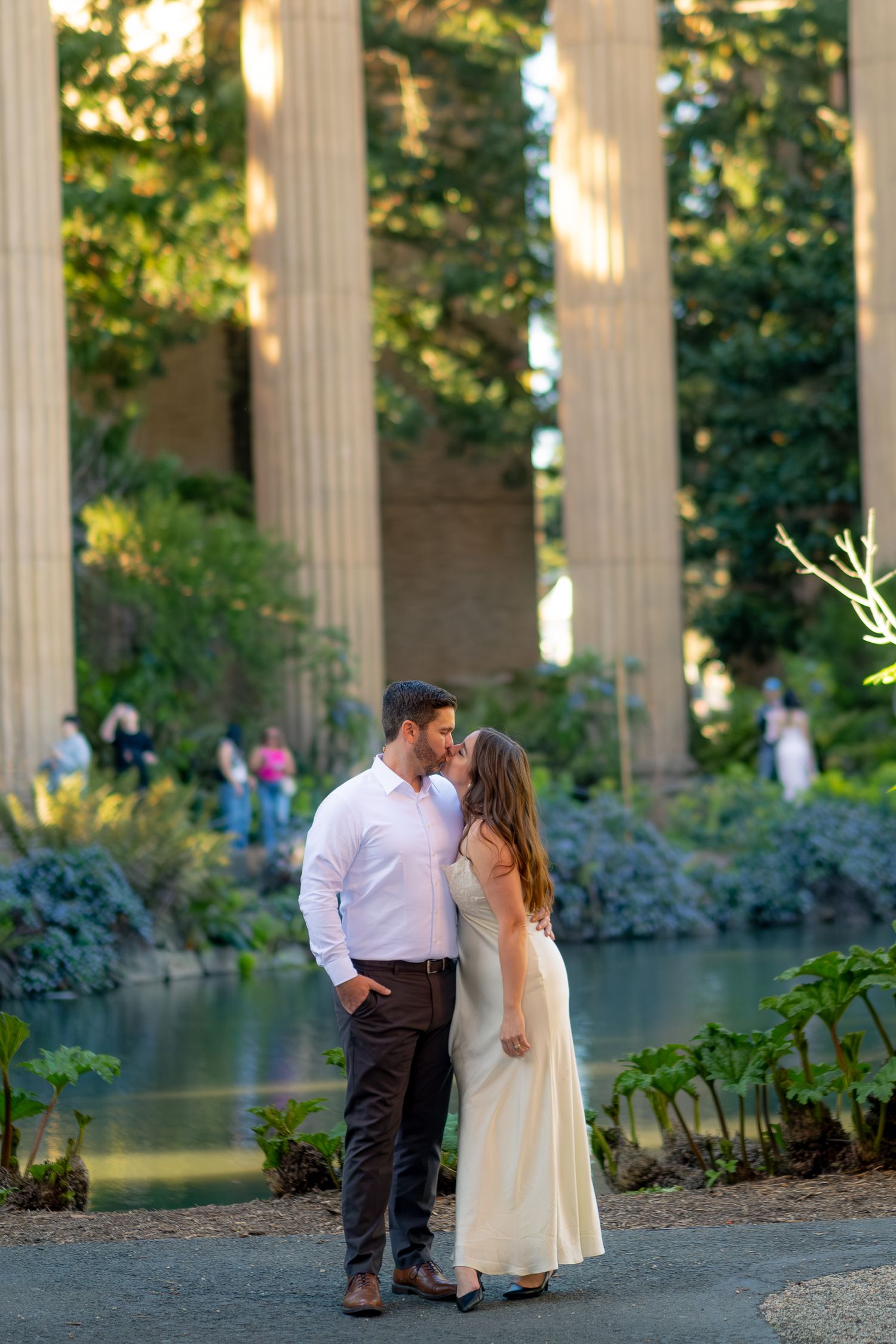 Engaged couple sharing a romantic moment near a reflective pond with towering columns behind them.