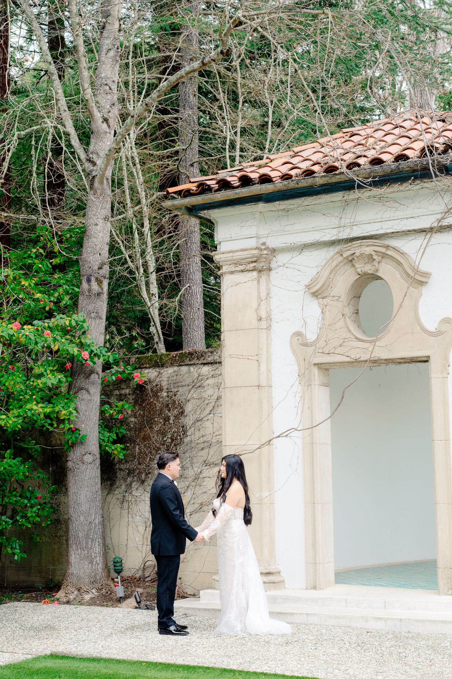 A bride and groom hold hands in a romantic outdoor setting beside an elegant white pavilion with a terracotta roof.