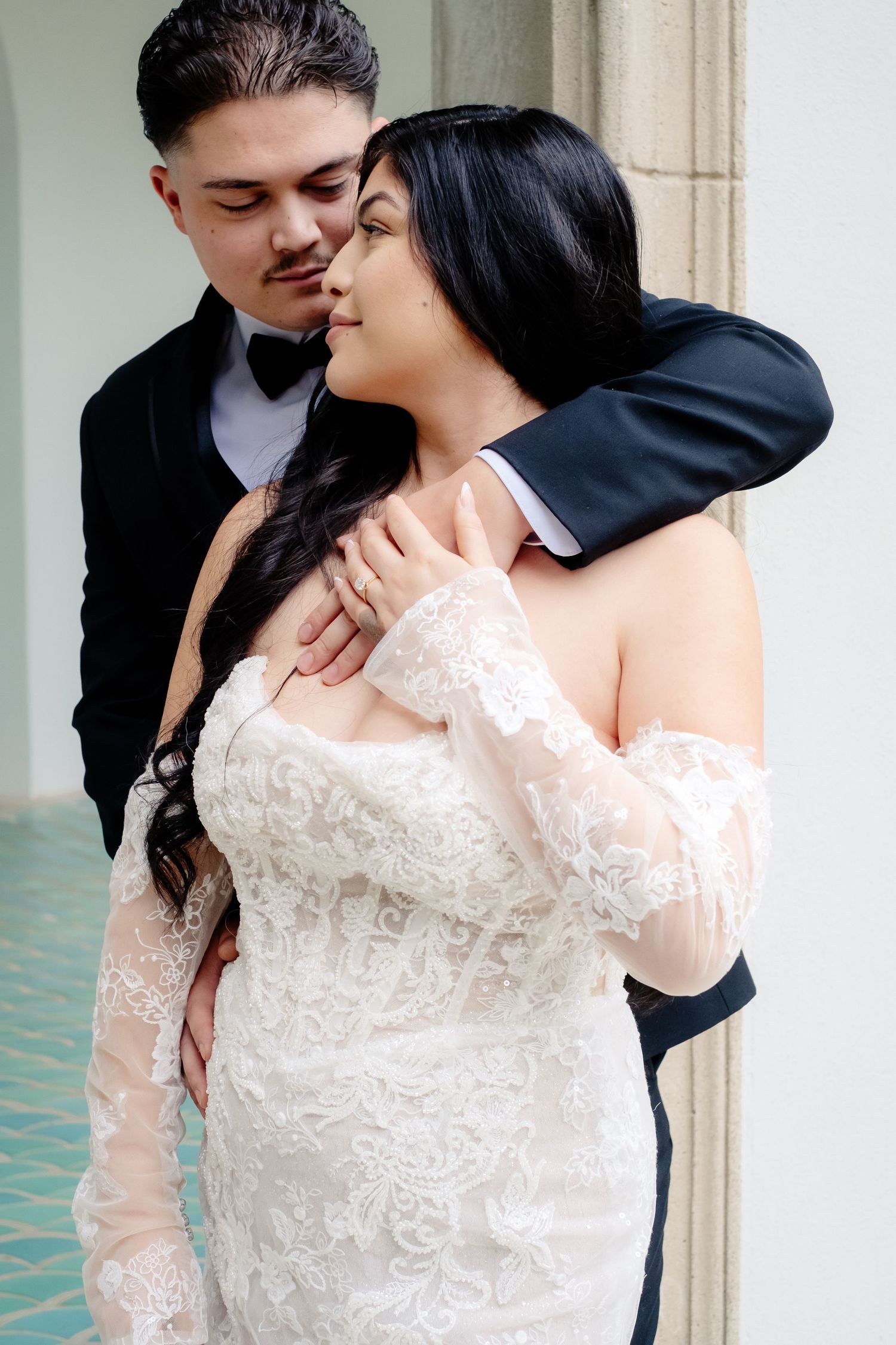 A bride in lace off-shoulder gown embraces her groom in a black tuxedo during romantic wedding portraits.