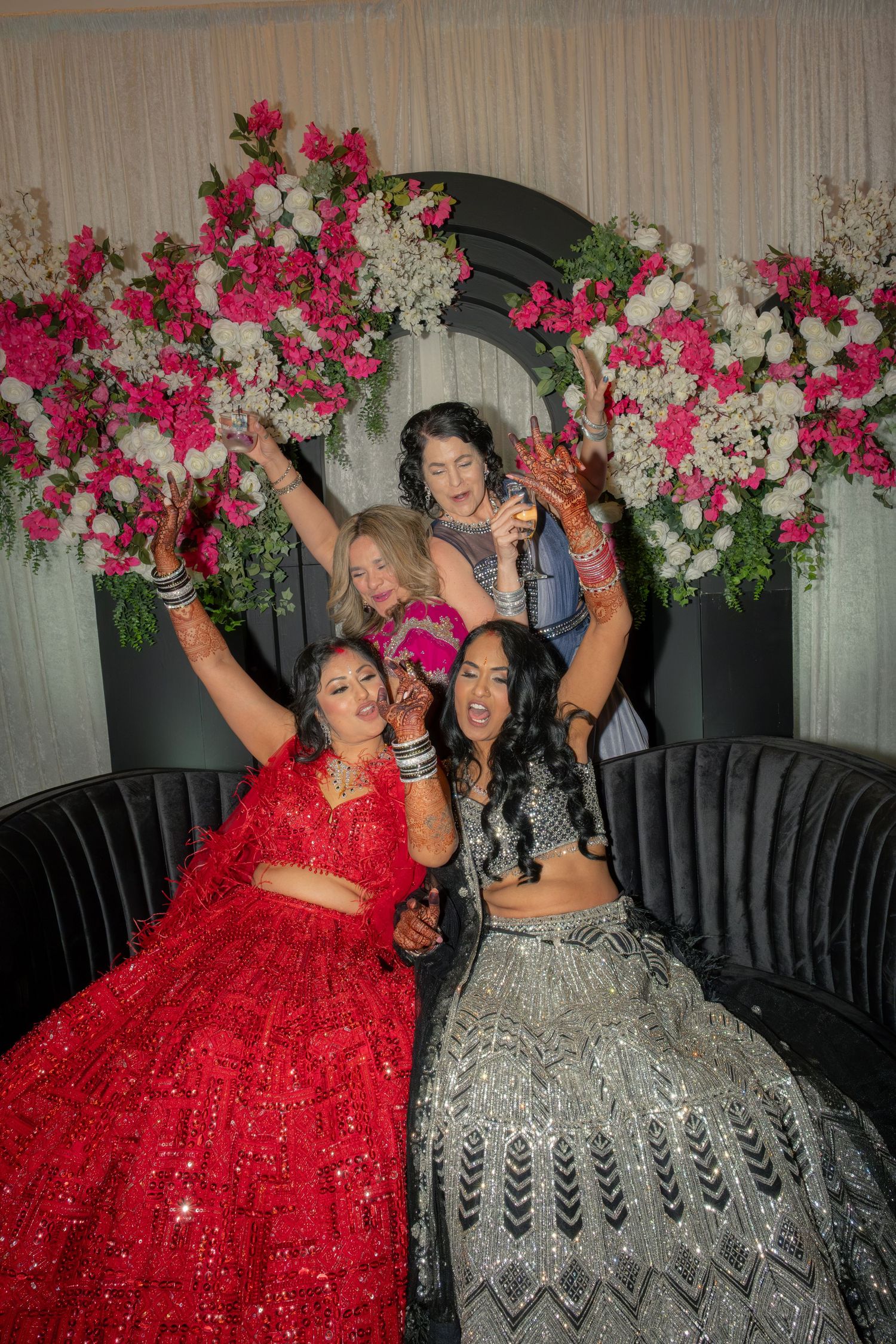 Four women celebrating at Indian wedding with pink and white floral backdrop, wearing red and silver lehengas.