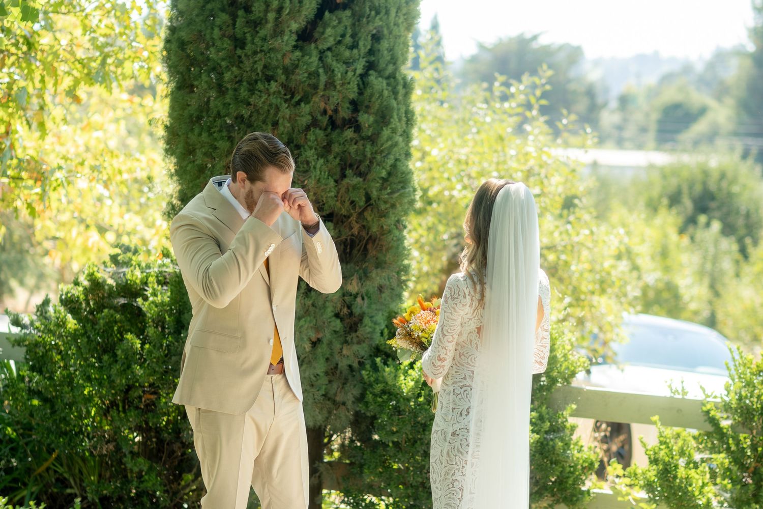 Emotional groom wiping tears during first look with bride in white veil holding bouquet at outdoor wedding.