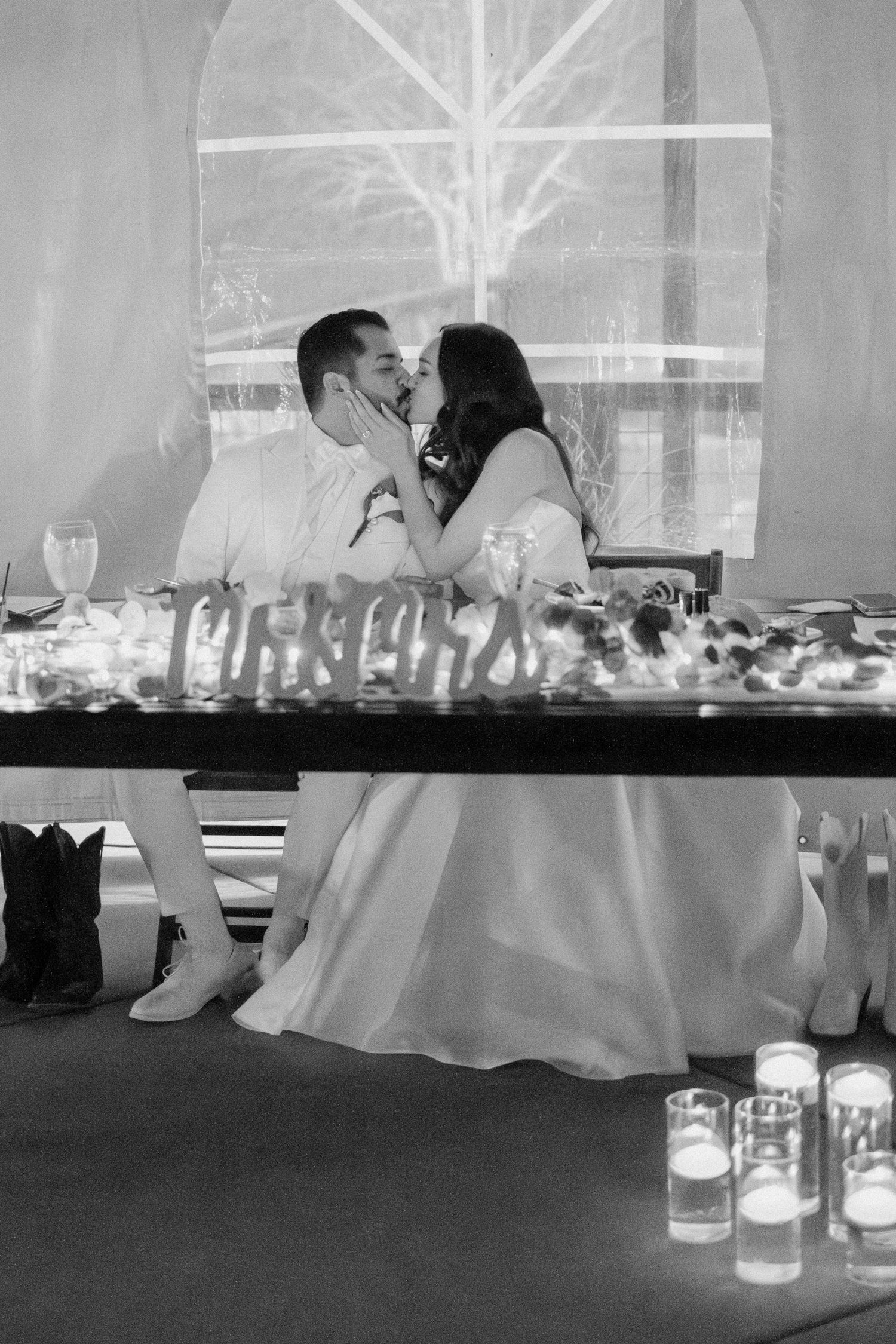 Newlyweds share a romantic kiss at their decorated sweetheart table with candles in black and white.