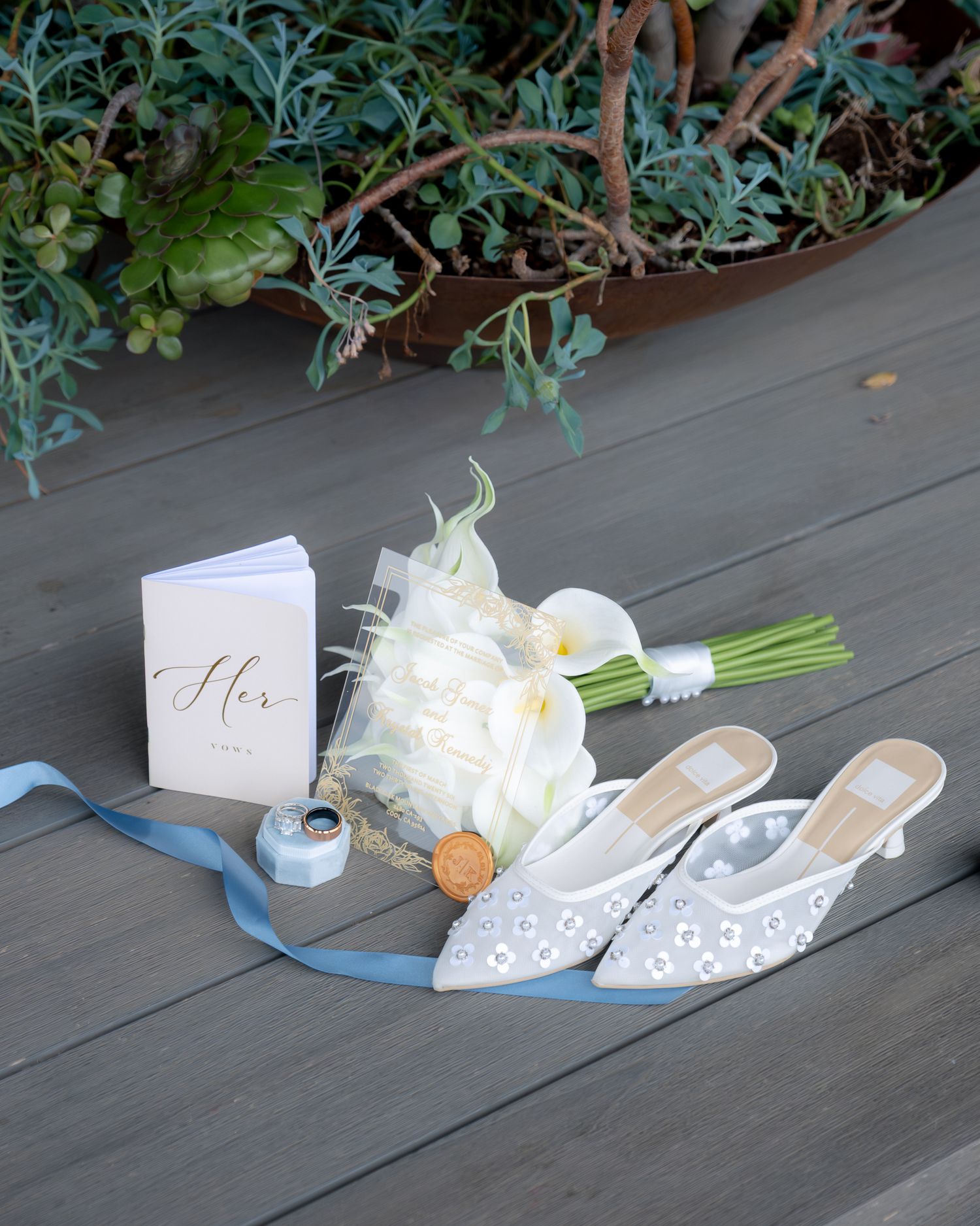 Elegant bridal flat lay with white calla lily bouquet, pearl heels, vows book, and blue ribbon on gray wood deck.