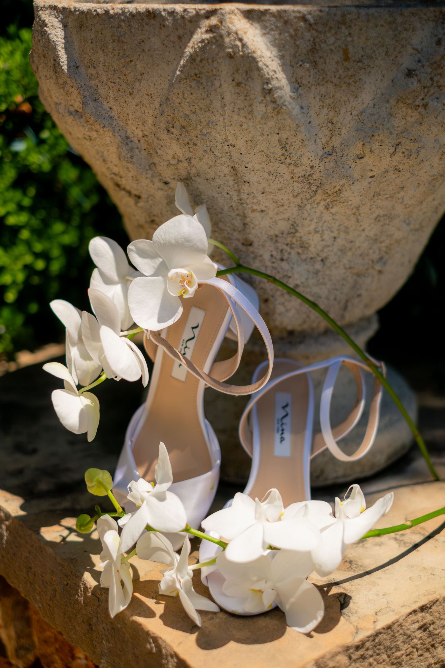 White bridal heels adorned with delicate white orchids displayed on a stone surface outdoors.