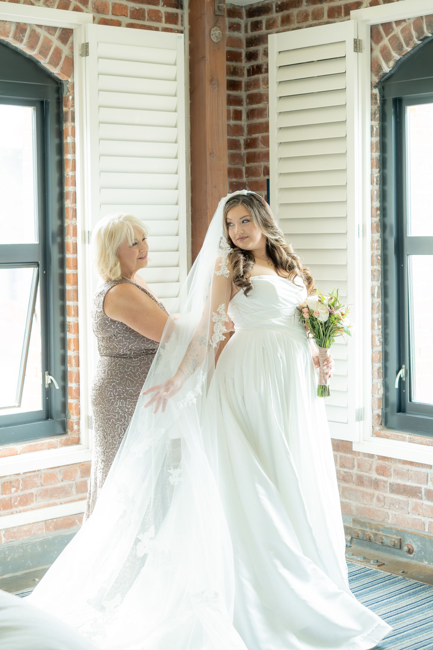 Mother helps bride adjust her lace veil in a brick-walled bridal suite before the wedding ceremony.