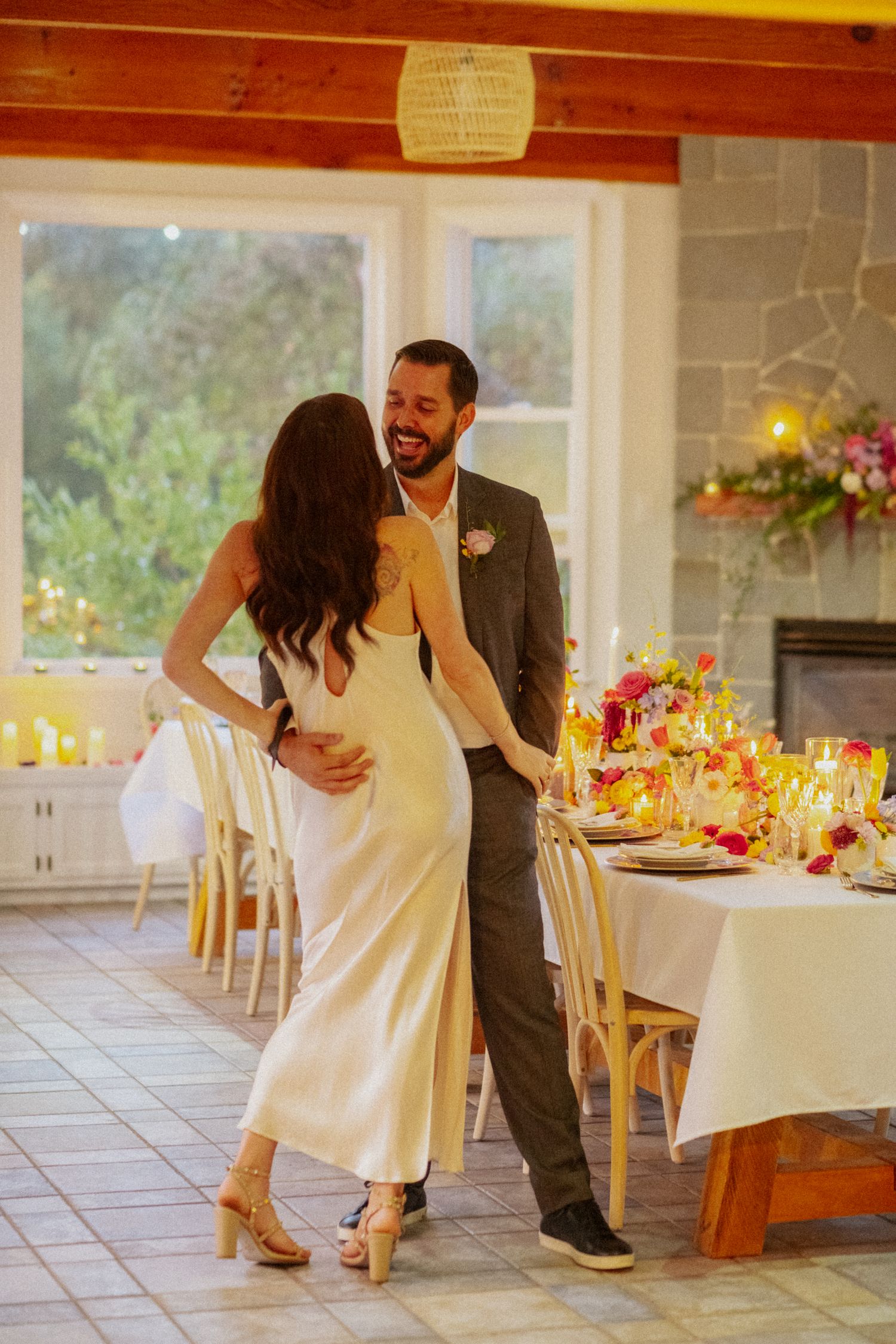 Bride in white satin dress and groom in gray suit share a joyful dance beside an elegantly decorated wedding reception table.